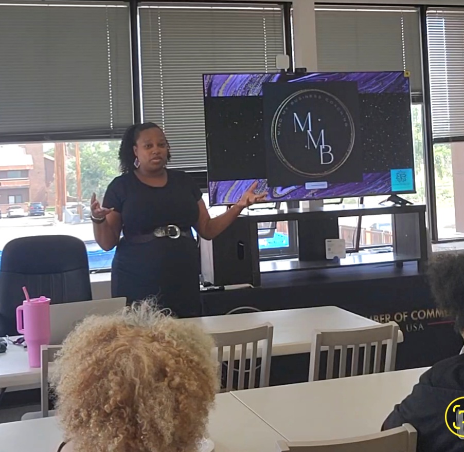 Woman presenting in a room with large windows, a TV screen displaying a logo, and seated audience members.