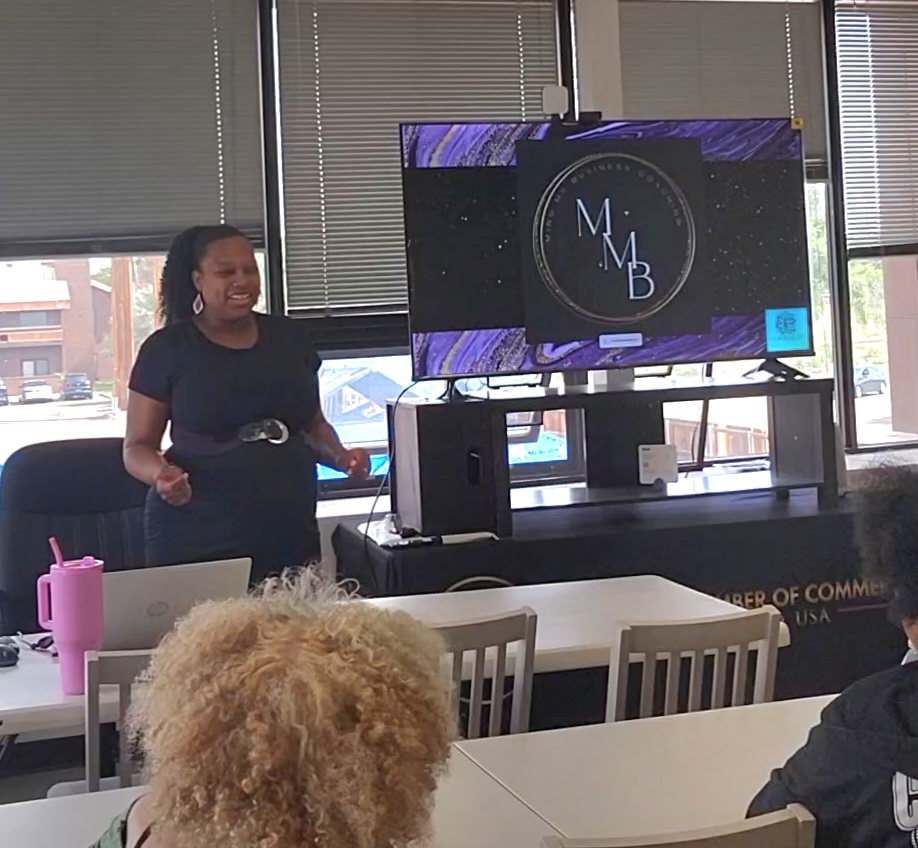 Woman standing and smiling in front of a large screen displaying a logo with the letters M, M, B. People seated at tables in a room with windows.