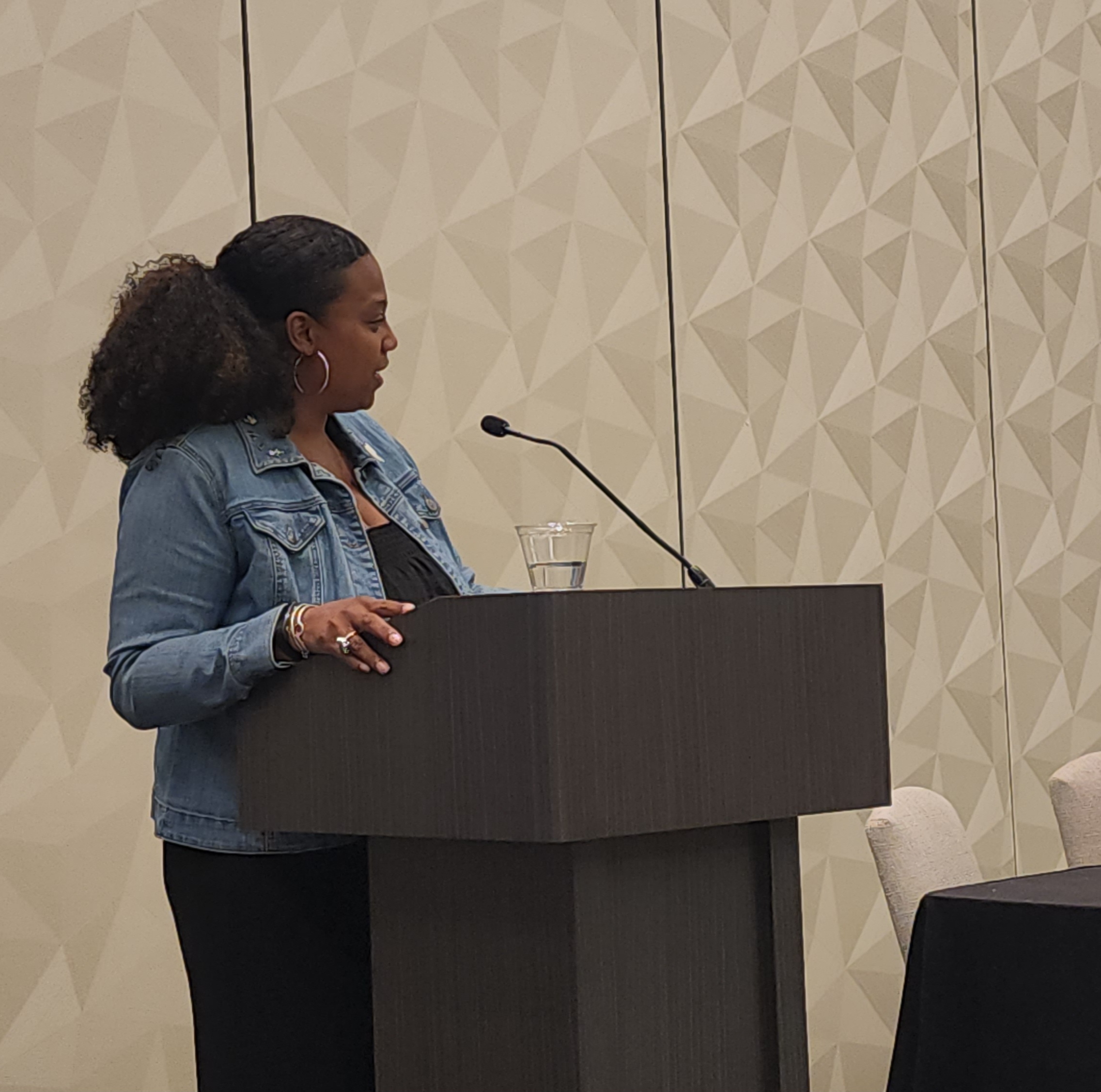 Woman standing at a podium speaking into a microphone, with a glass of water nearby, in a conference room.