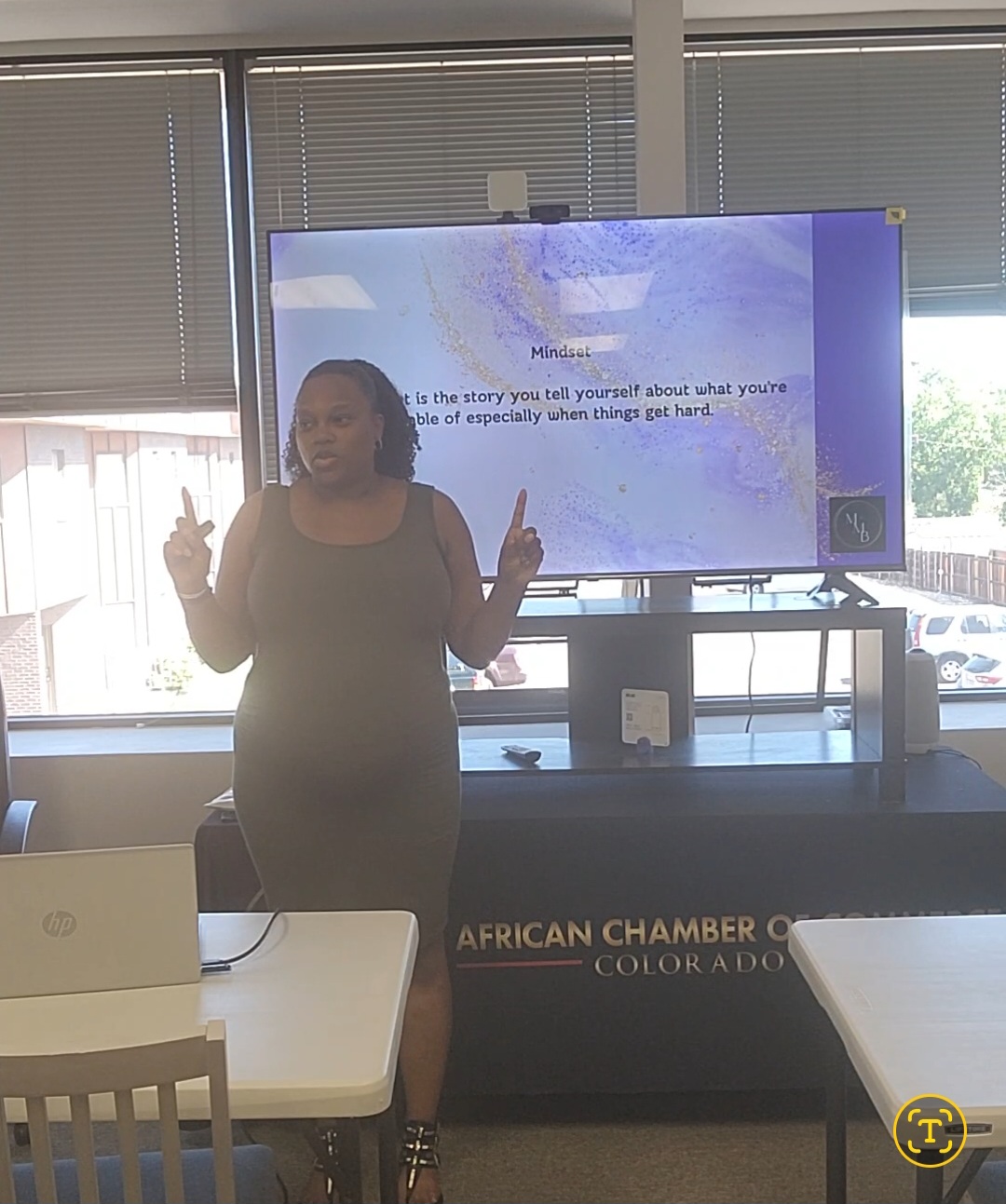 Woman standing in front of a screen, gesturing with both hands, in a room with windows and tables, African Chamber of Commerce sign visible.