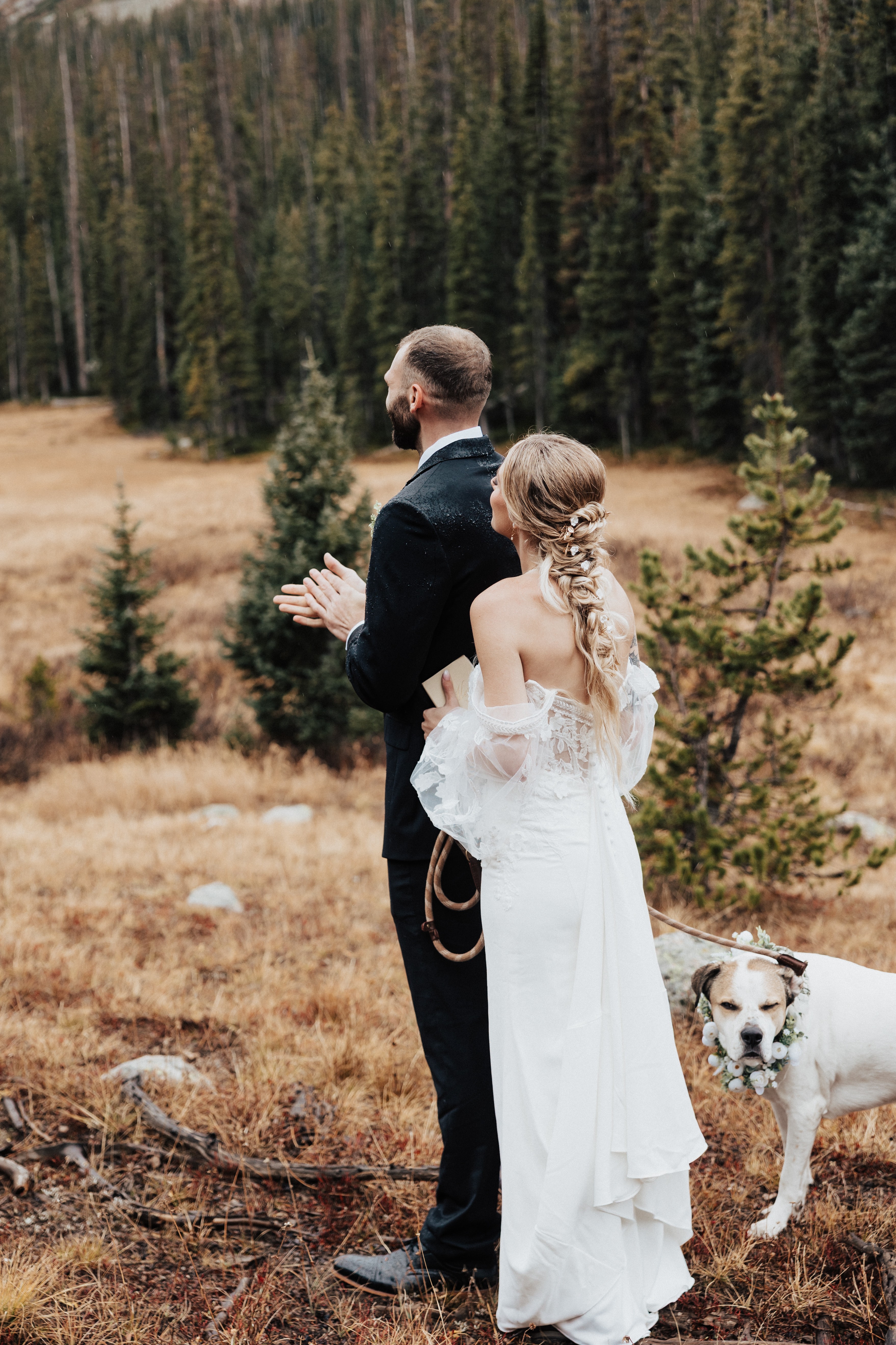 A bride and groom stand outdoors in a field with trees, facing away, with a dog nearby, during a wedding ceremony.