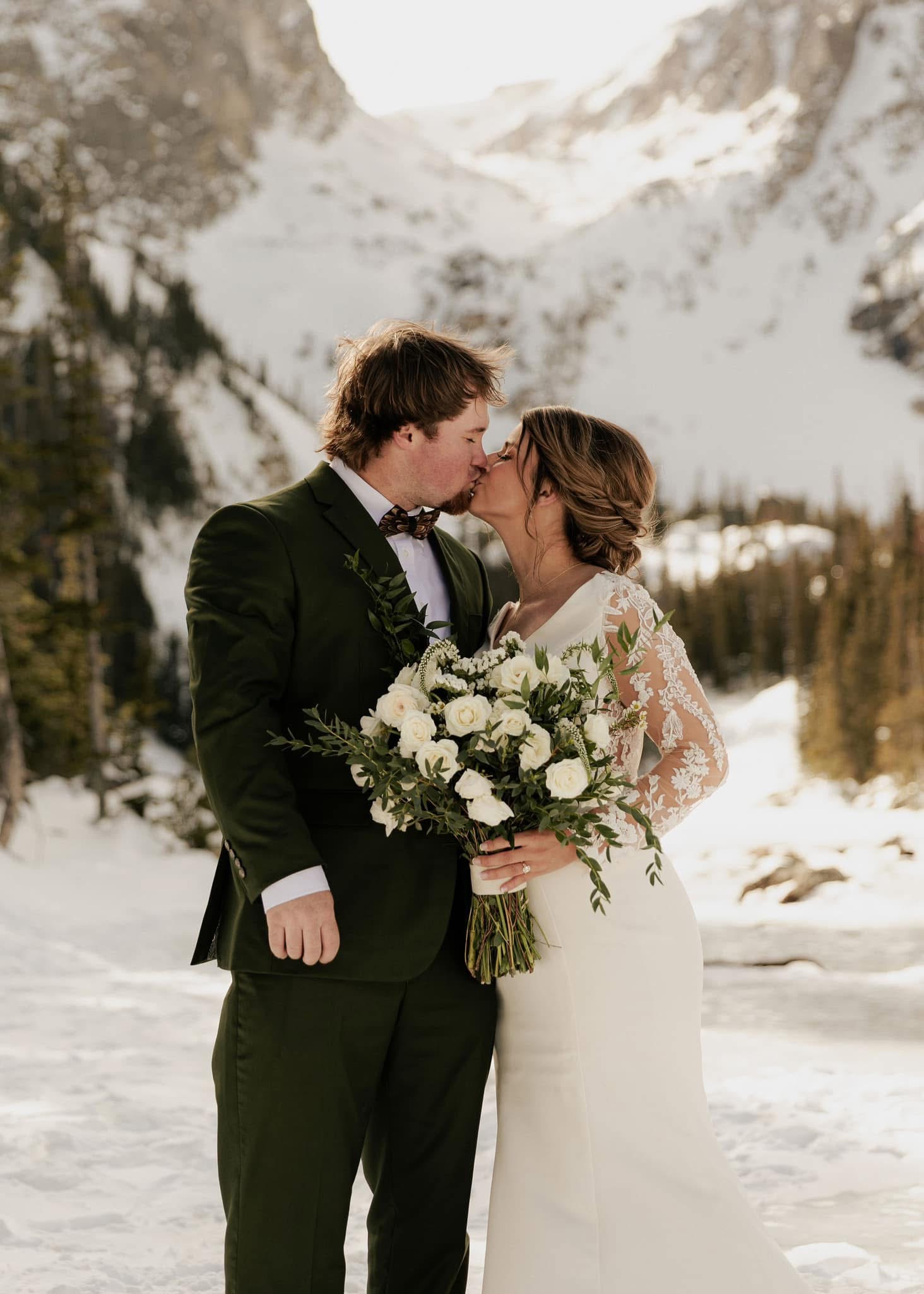 Couple in wedding attire kissing outdoors in snowy mountain landscape, woman holding a large bouquet of white flowers.