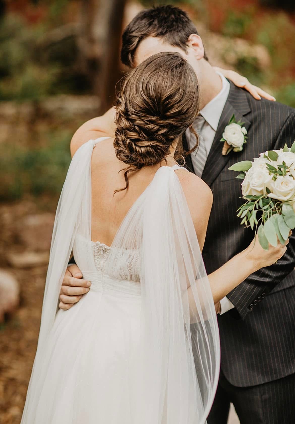 A bride and groom embrace outdoors, the bride holding a bouquet, with trees and autumn foliage in the background.