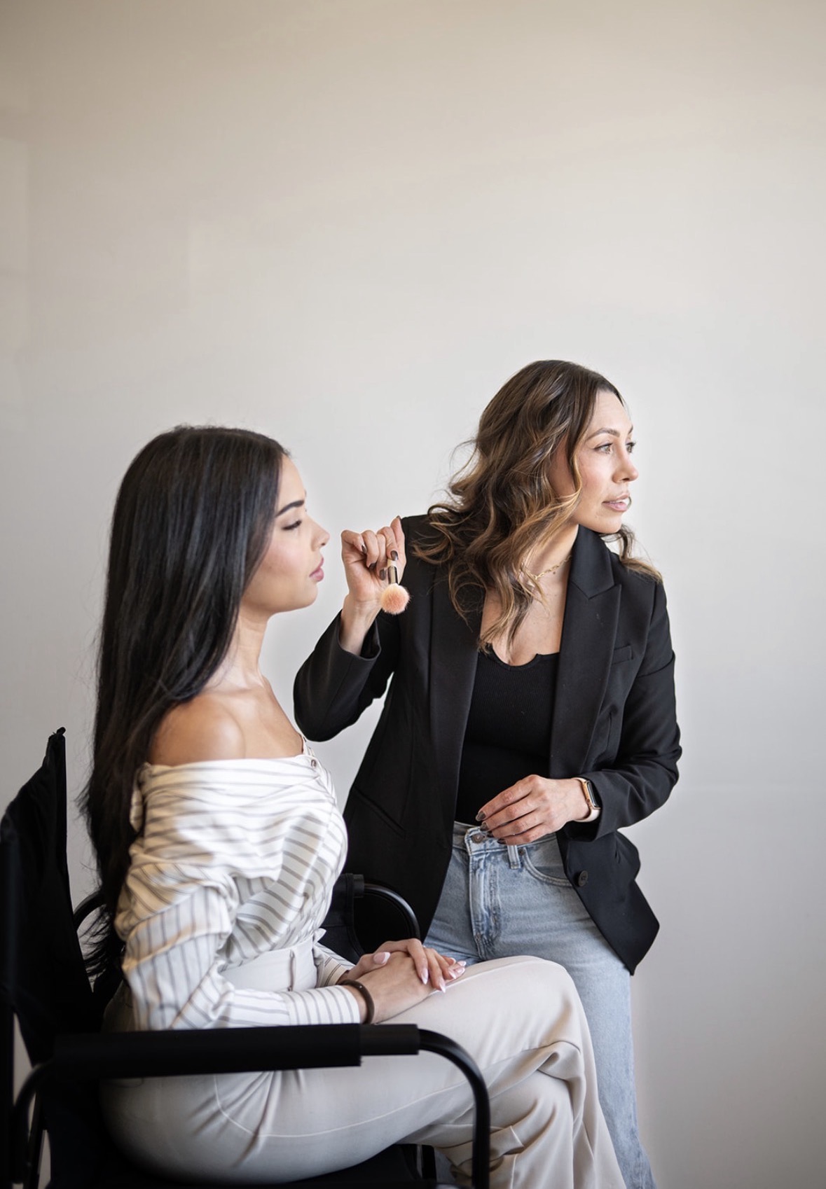 Two women sit and talk, one with long dark hair and the other with shoulder-length brown hair, against a plain wall.