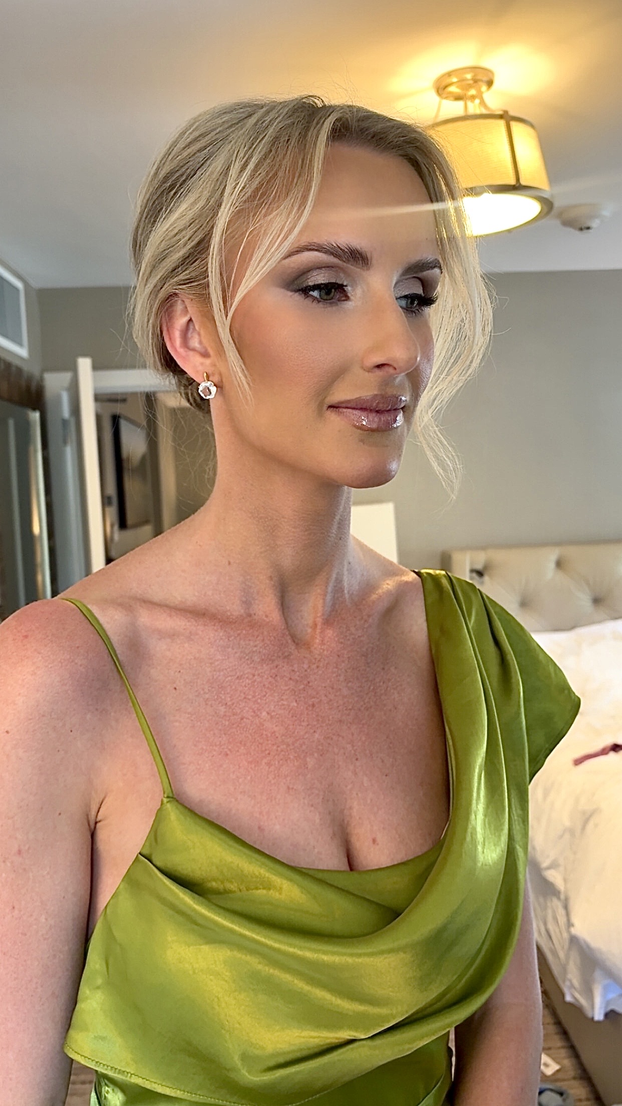 Woman with blonde hair in an updo, wearing a green satin dress with thin straps, in a bedroom setting.