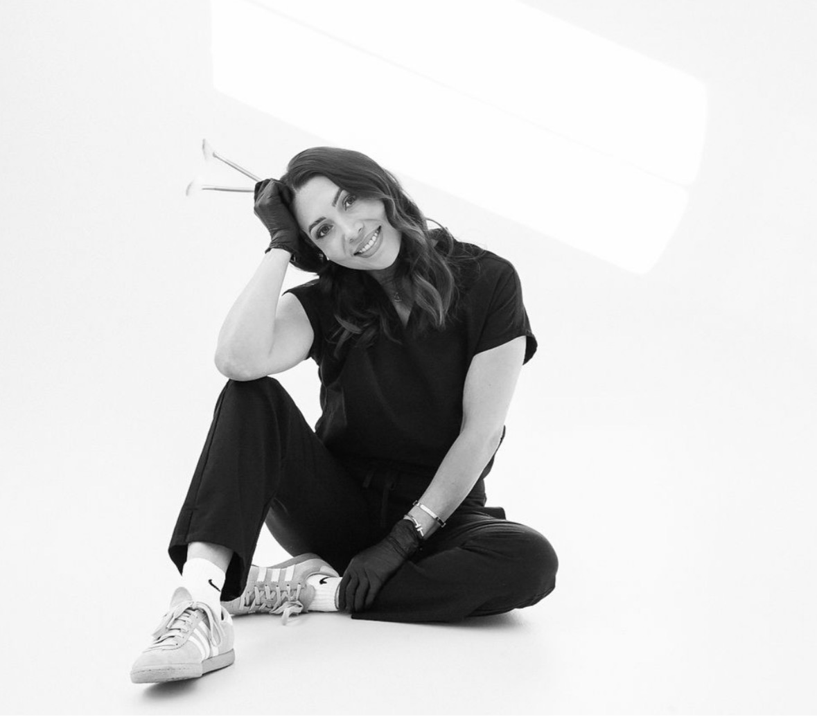 Woman sitting on the floor, smiling, with one hand on her head, wearing a black outfit and sneakers.