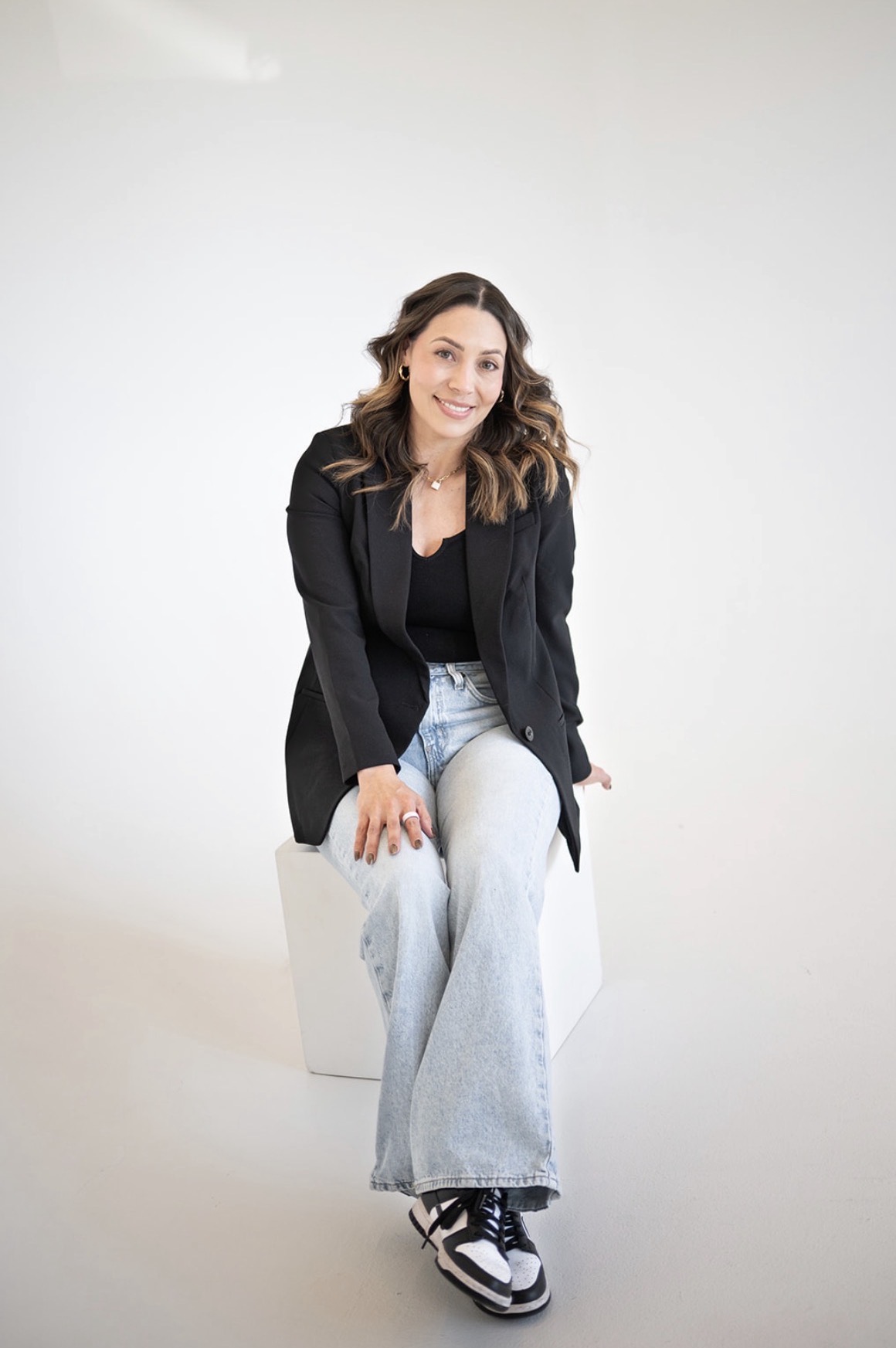 Woman sitting on a white block, smiling, wearing a black blazer, black top, light blue jeans, and sneakers, against a plain white background.