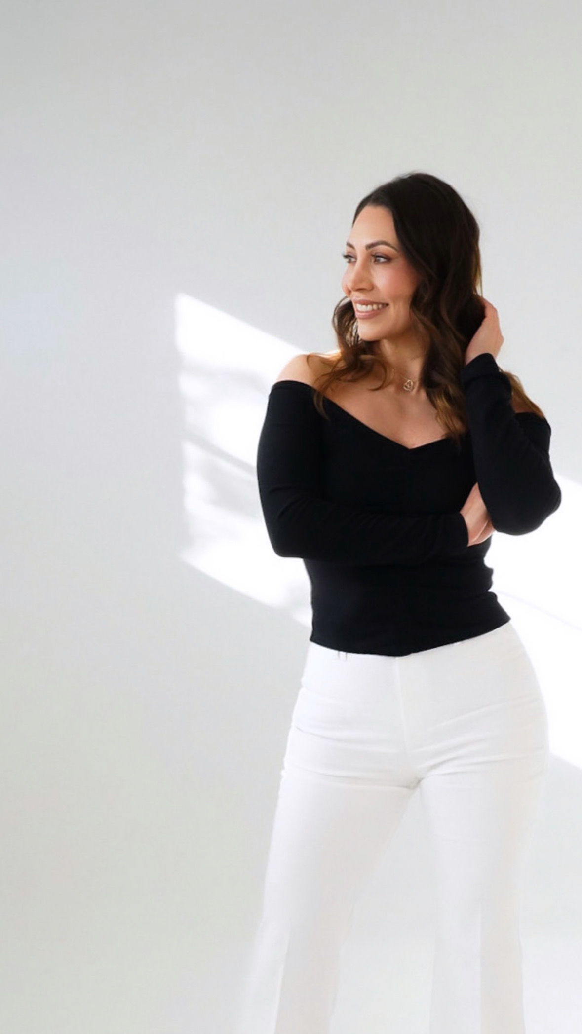 Woman with dark hair smiling, wearing a black top and white pants, standing against a plain light background.