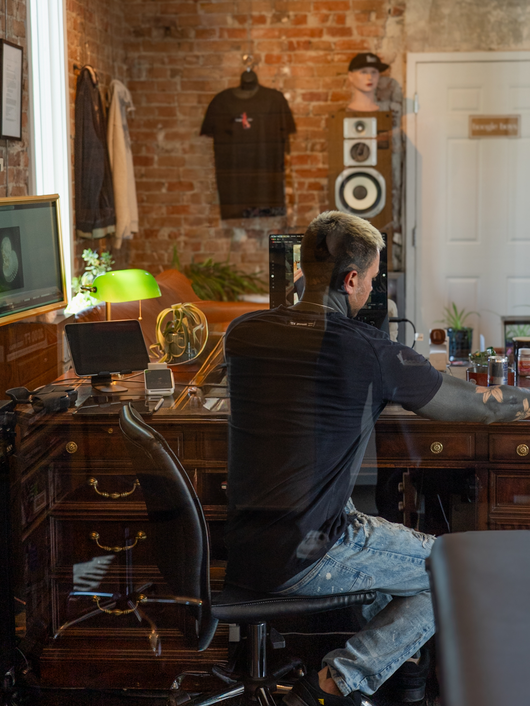 Man sitting at a wooden desk in a room with brick walls, speakers, and clothing hanging on the wall.