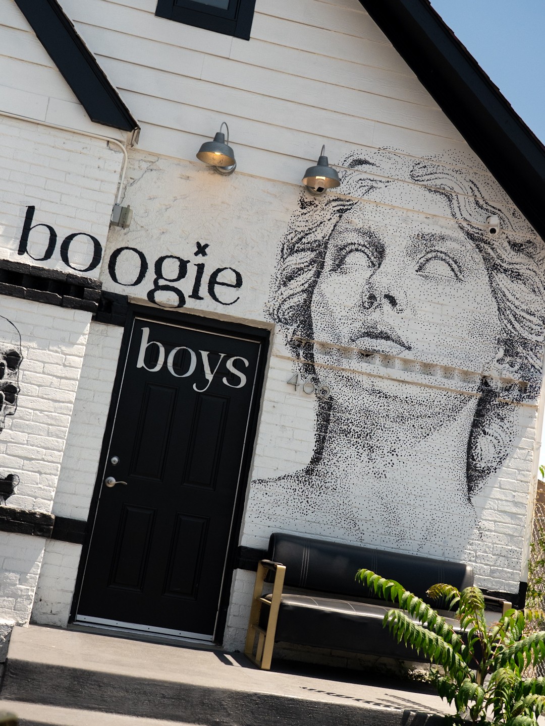 White brick building with a large mural of a woman's face, black door labeled 'boys', and a bench outside.