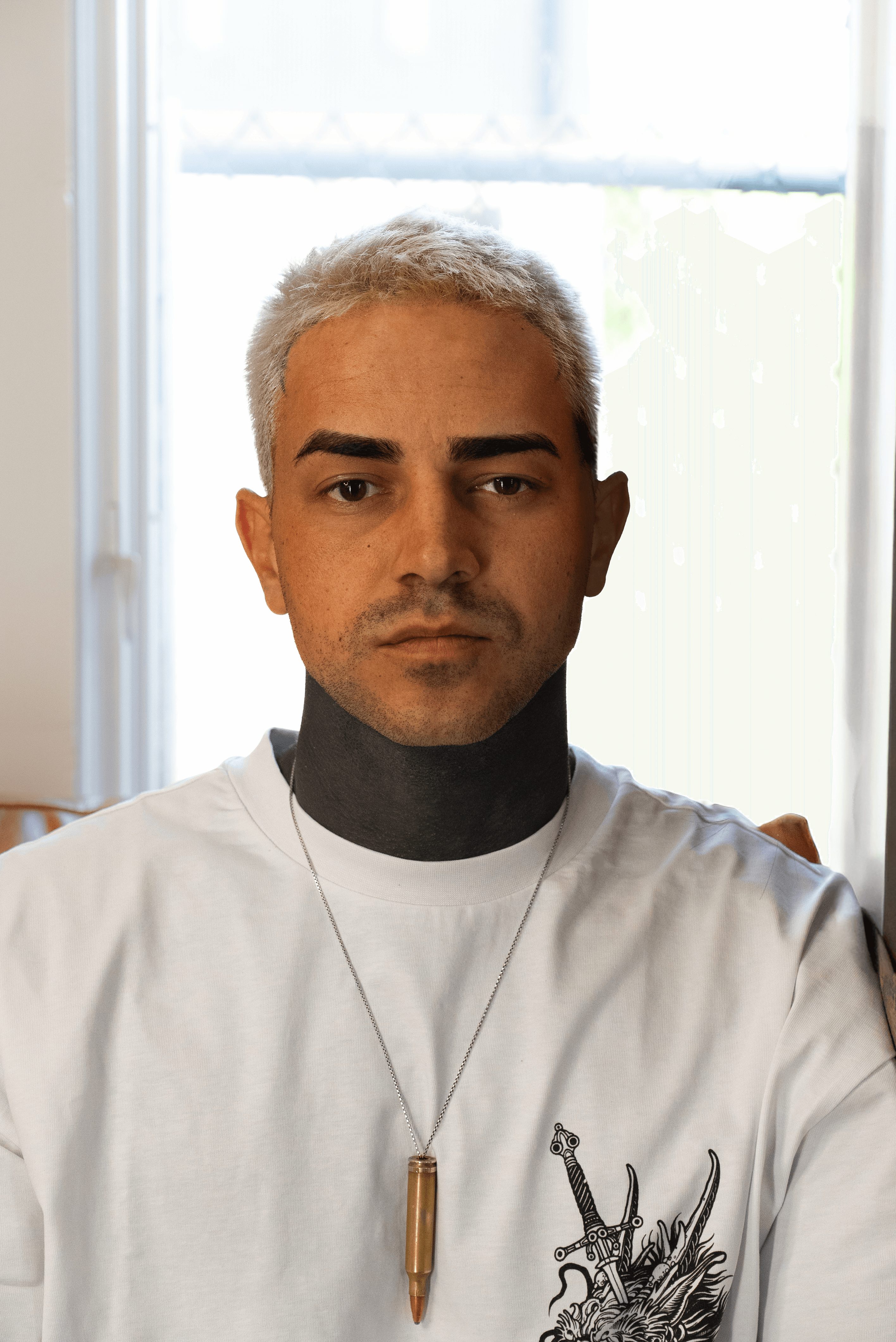 Young man with short, light-colored hair, wearing a white shirt with a black neck covering, and a necklace with a pendant, standing indoors near a window.