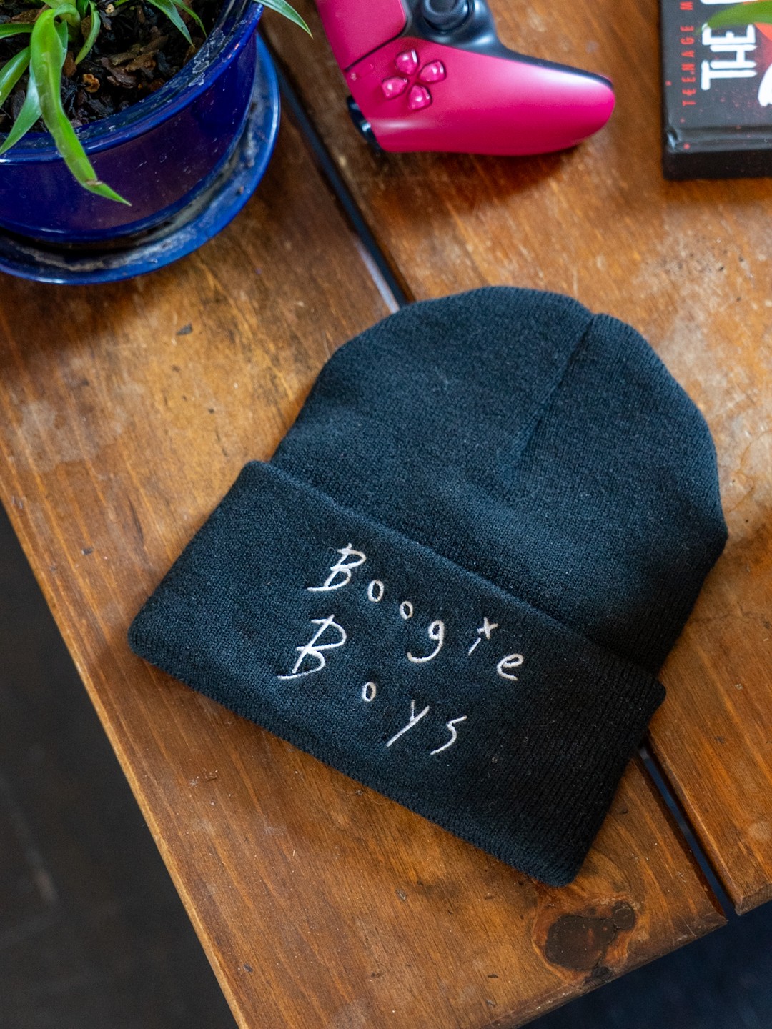 Black beanie with white embroidered text reading 'Bogie Boys' on a wooden surface, with a pink game controller and a plant nearby.
