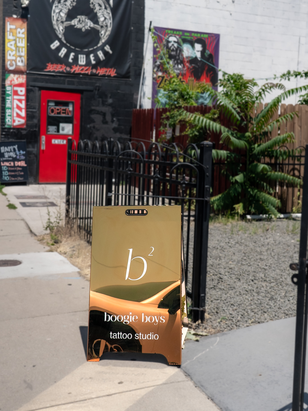 A sidewalk sign advertising boogie boys tattoo studio with a gold and black design, in front of a black fence and plants.