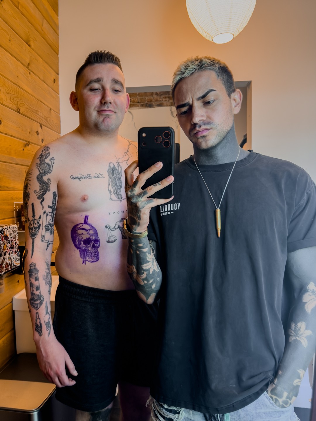 Two young men taking a mirror selfie indoors, one shirtless with tattoos, the other wearing a black T-shirt, both looking at the camera.