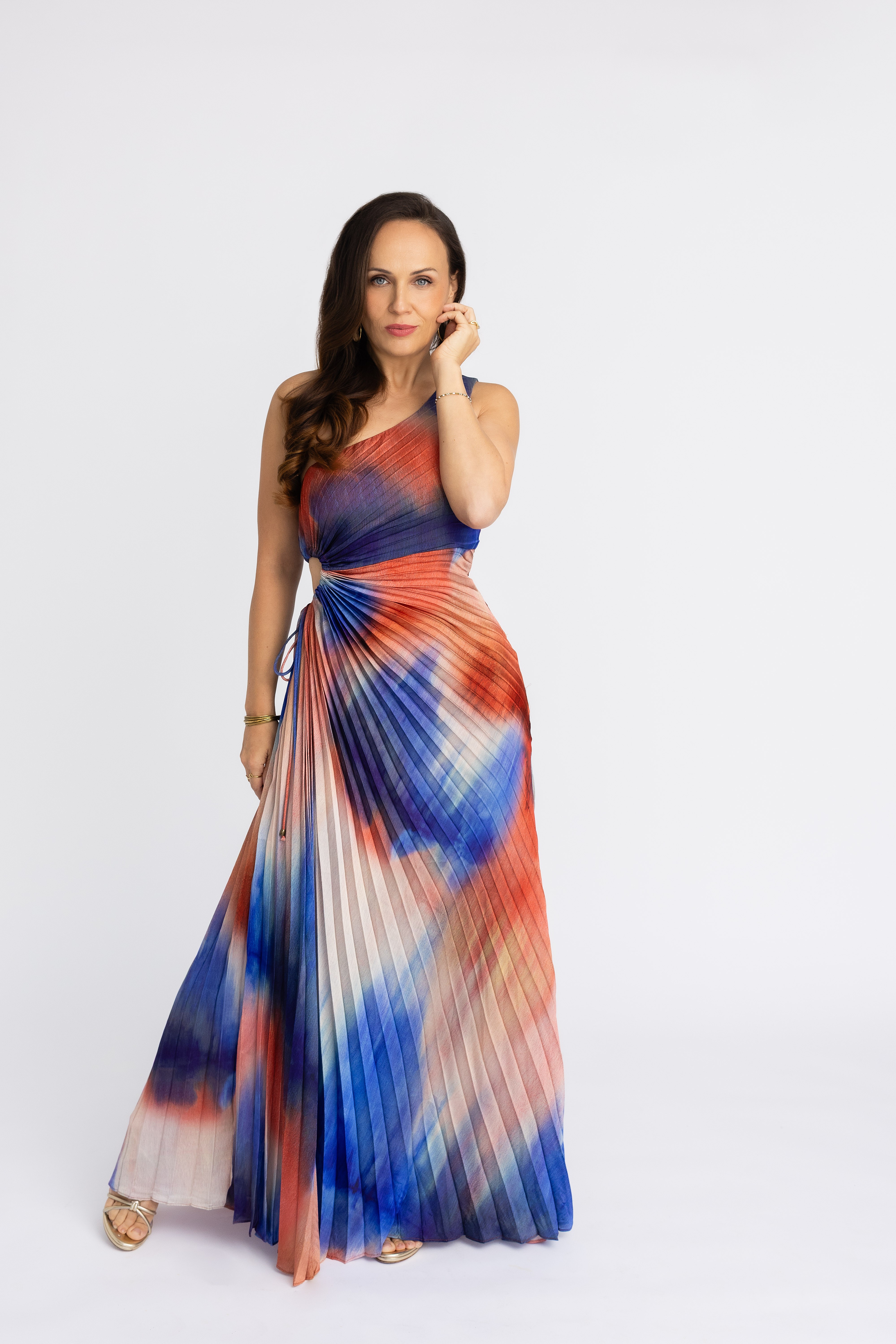 Woman in a colorful, pleated maxi dress with a tie-dye pattern, standing against a plain white background.