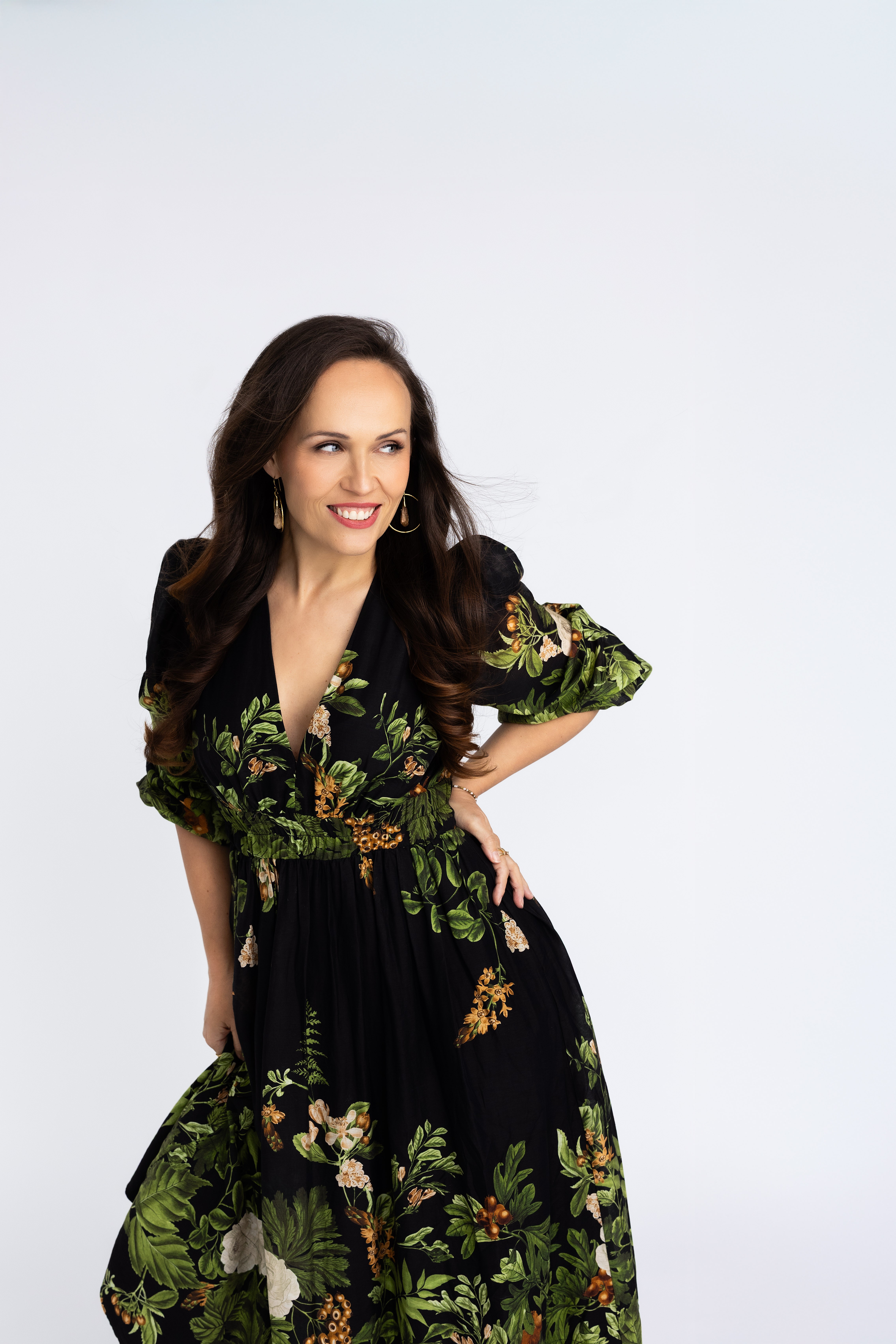 Woman with long dark hair wearing a black floral dress, smiling, with one hand on her hip, against a plain white background.