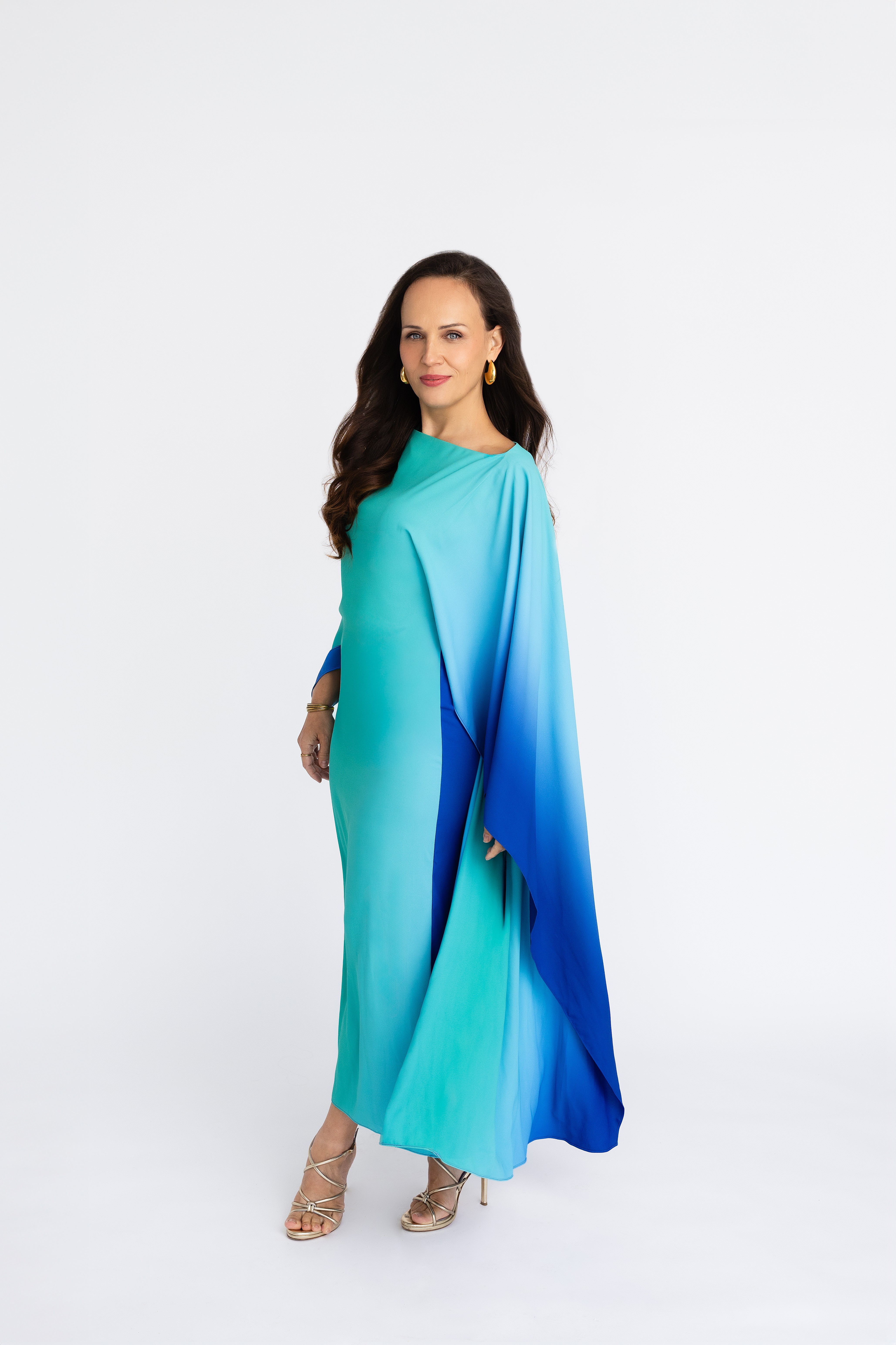 Woman in a flowing blue and green dress with long dark hair, standing against a white background.