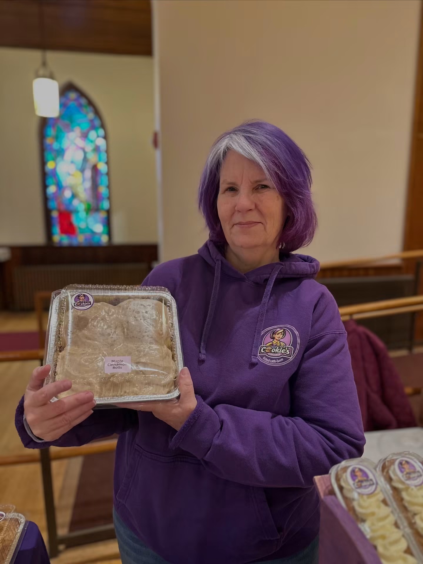 Woman with purple hair holding a cake in a church interior with stained glass window.