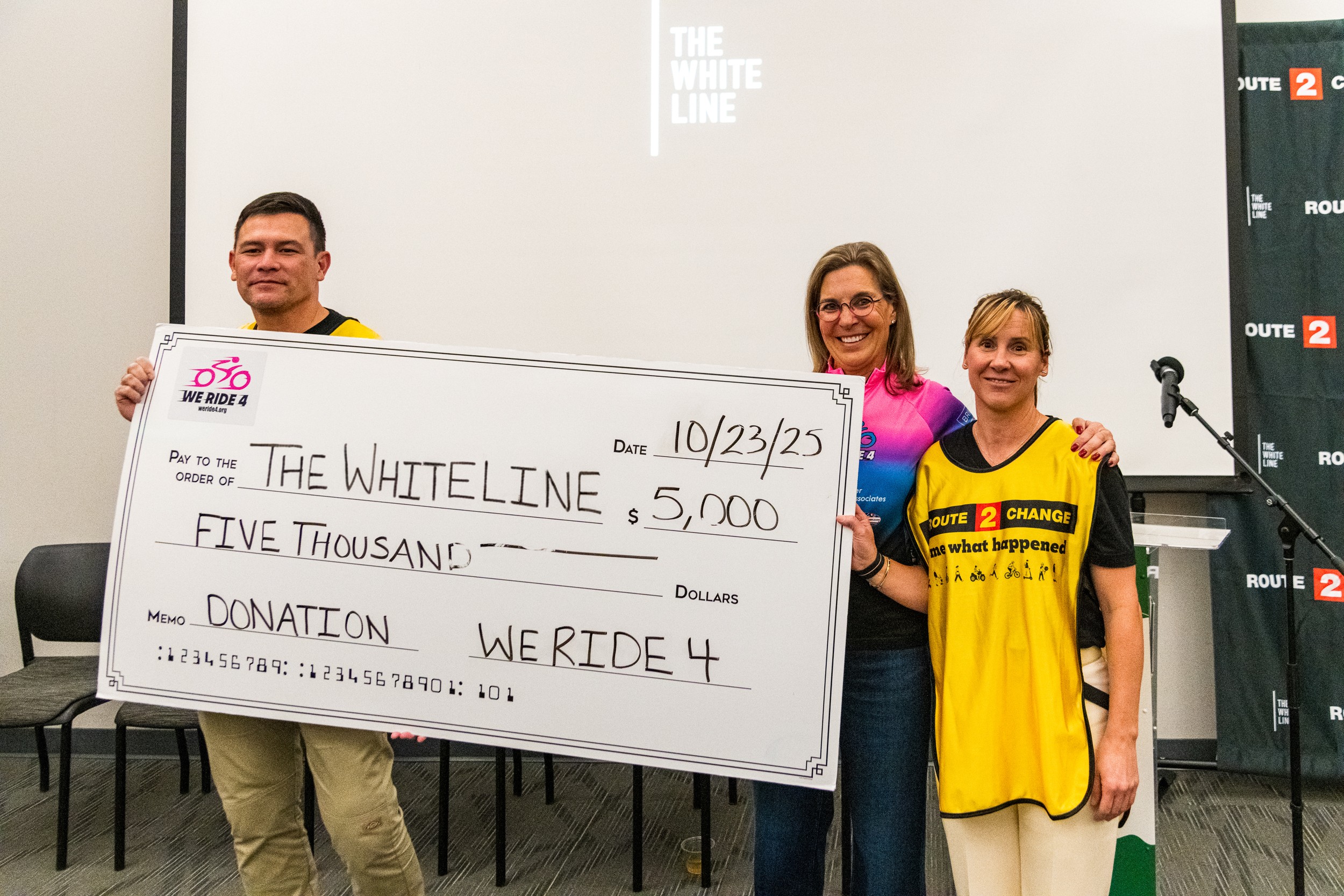 Three people standing in front of a screen holding a large check for five thousand dollars, smiling.