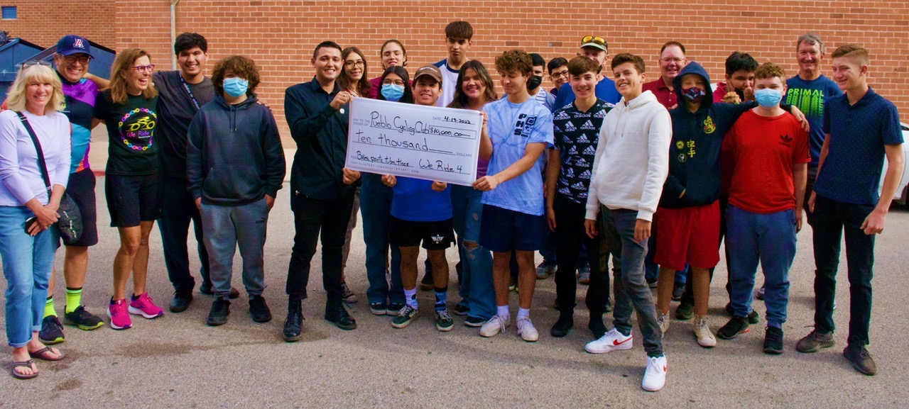 Group of people standing outdoors, holding a large check, smiling, in front of a brick wall.