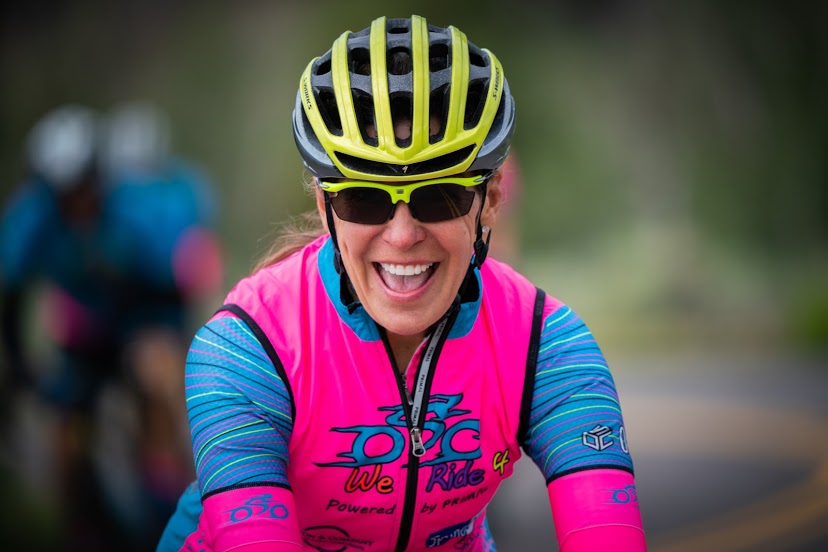 Smiling woman wearing a yellow helmet, sunglasses, and a pink and blue cycling jersey outdoors.