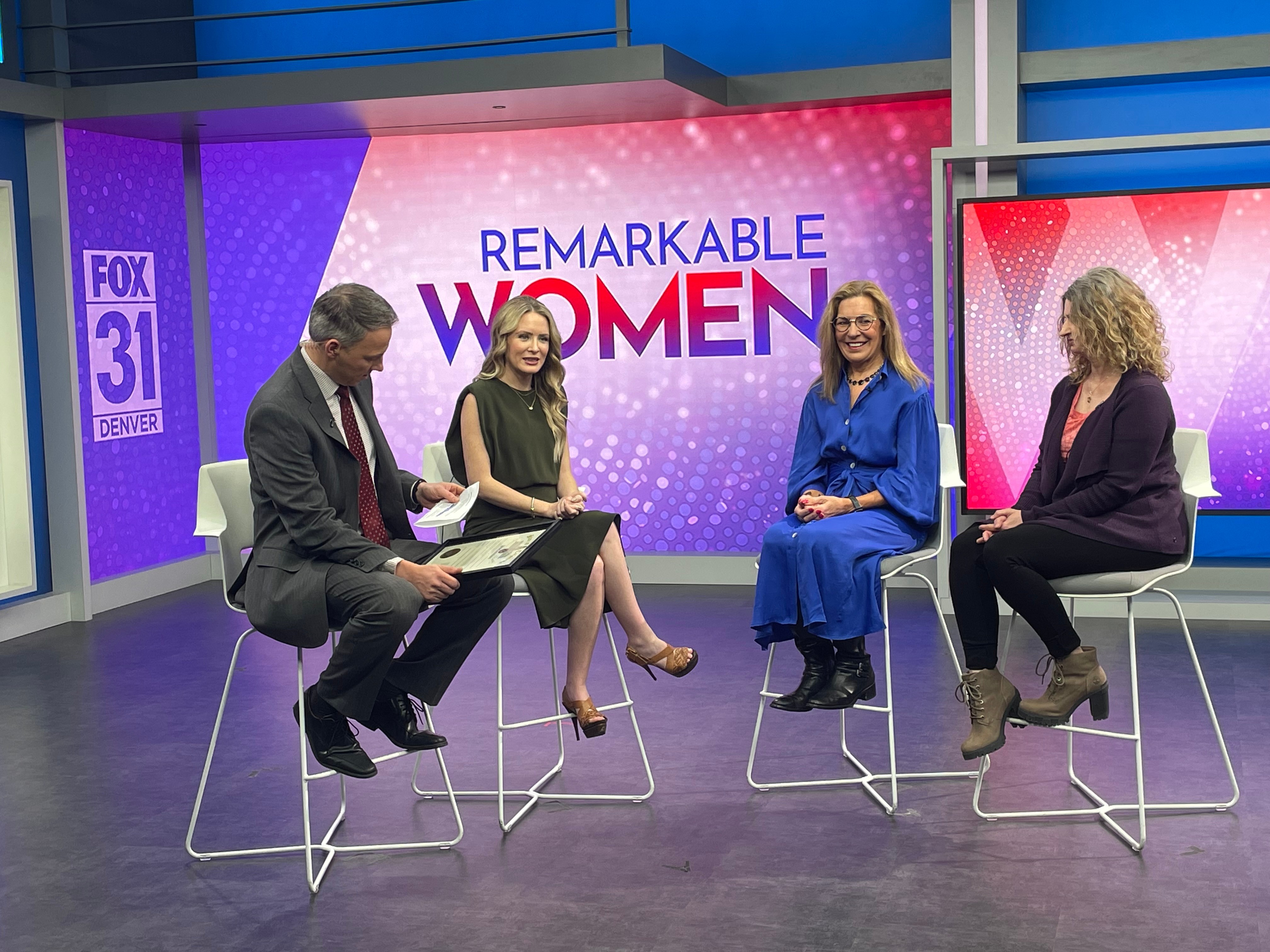 Four women and one man seated on a TV set with a large screen behind them displaying 'Remarkable Women' and 'FOX 31'.