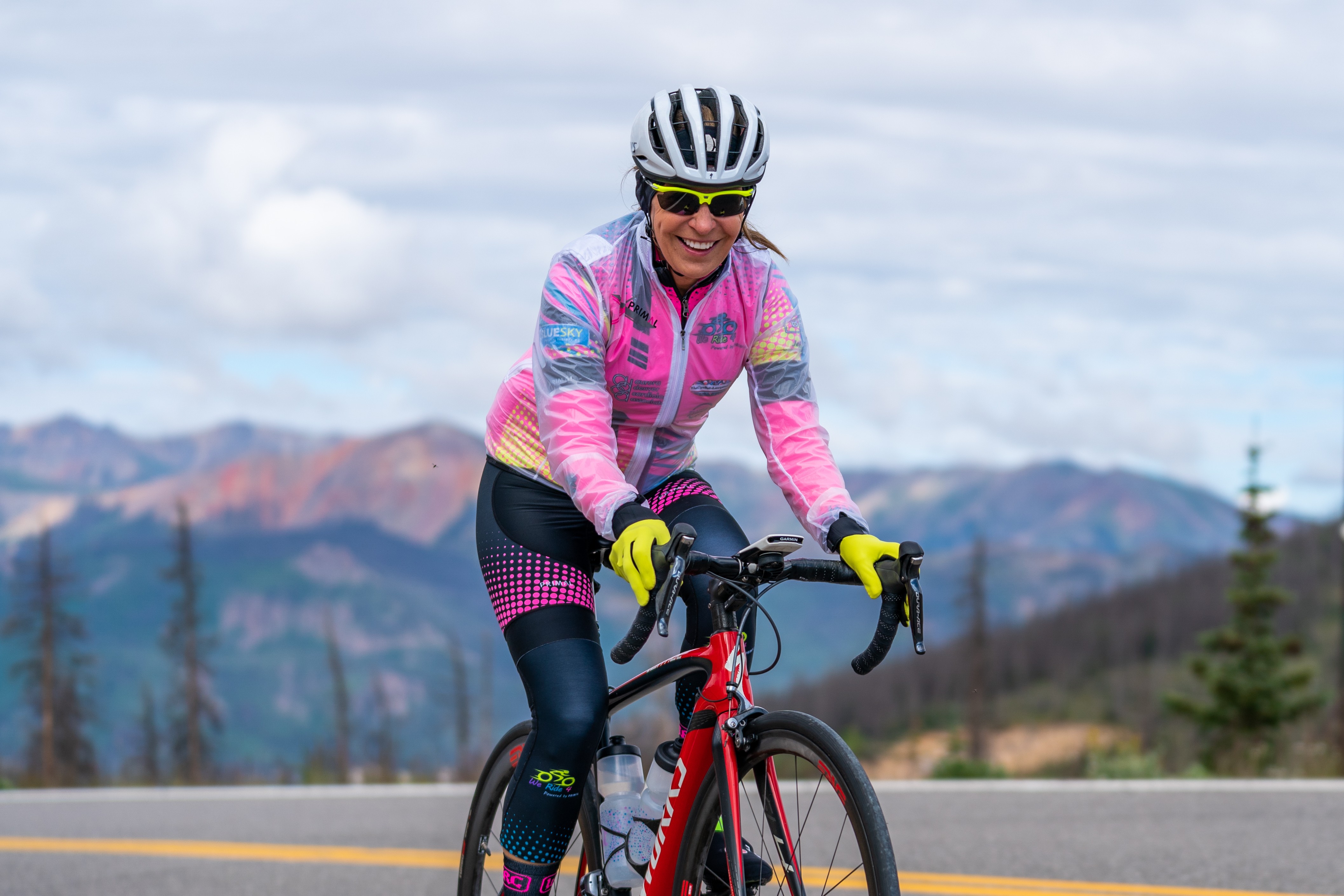 A woman in colorful cycling gear and helmet rides a red bike on a mountain road with mountains in the background.
