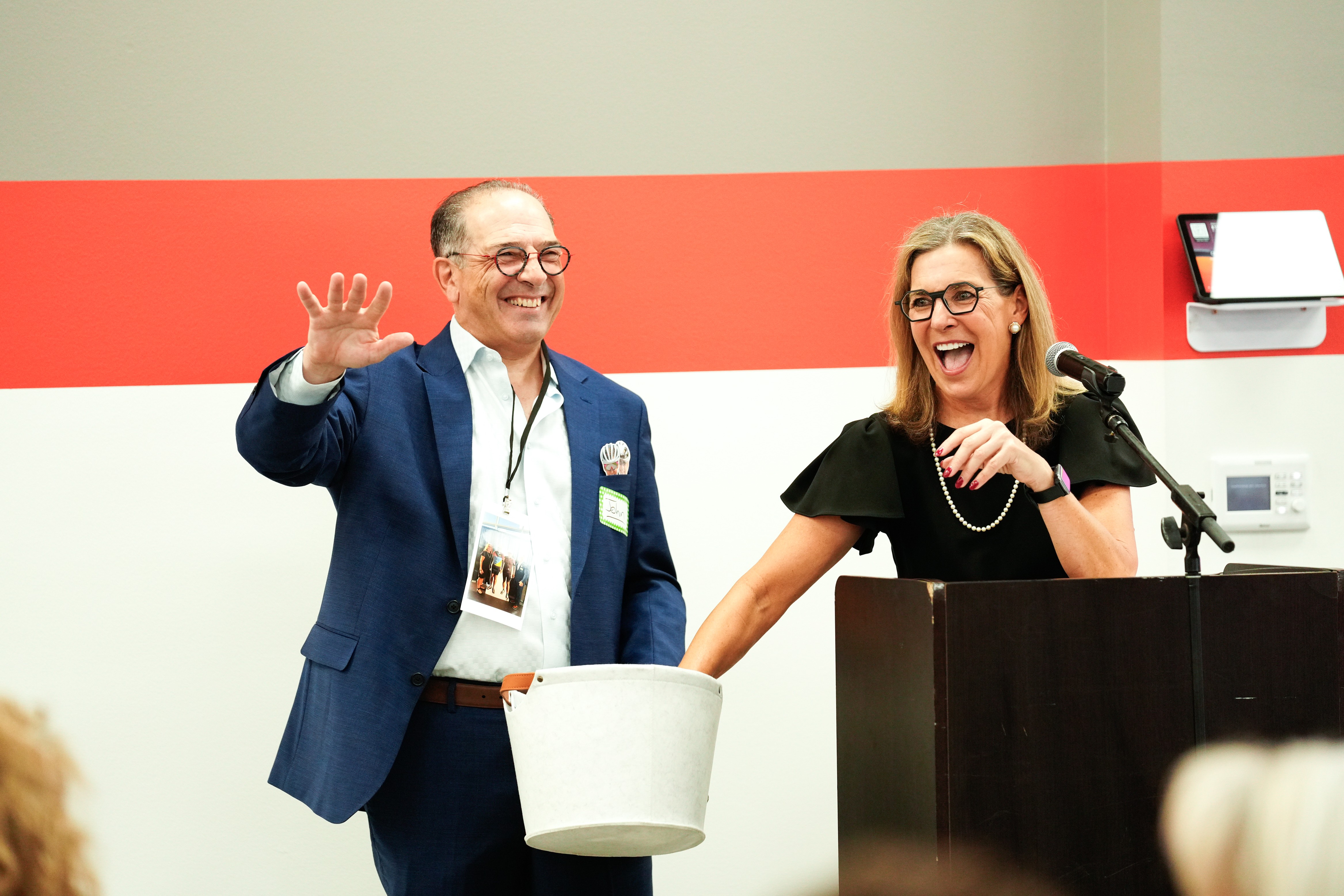 Two people smiling and laughing, one waving, standing behind a podium with a microphone, in a room with red and white walls.