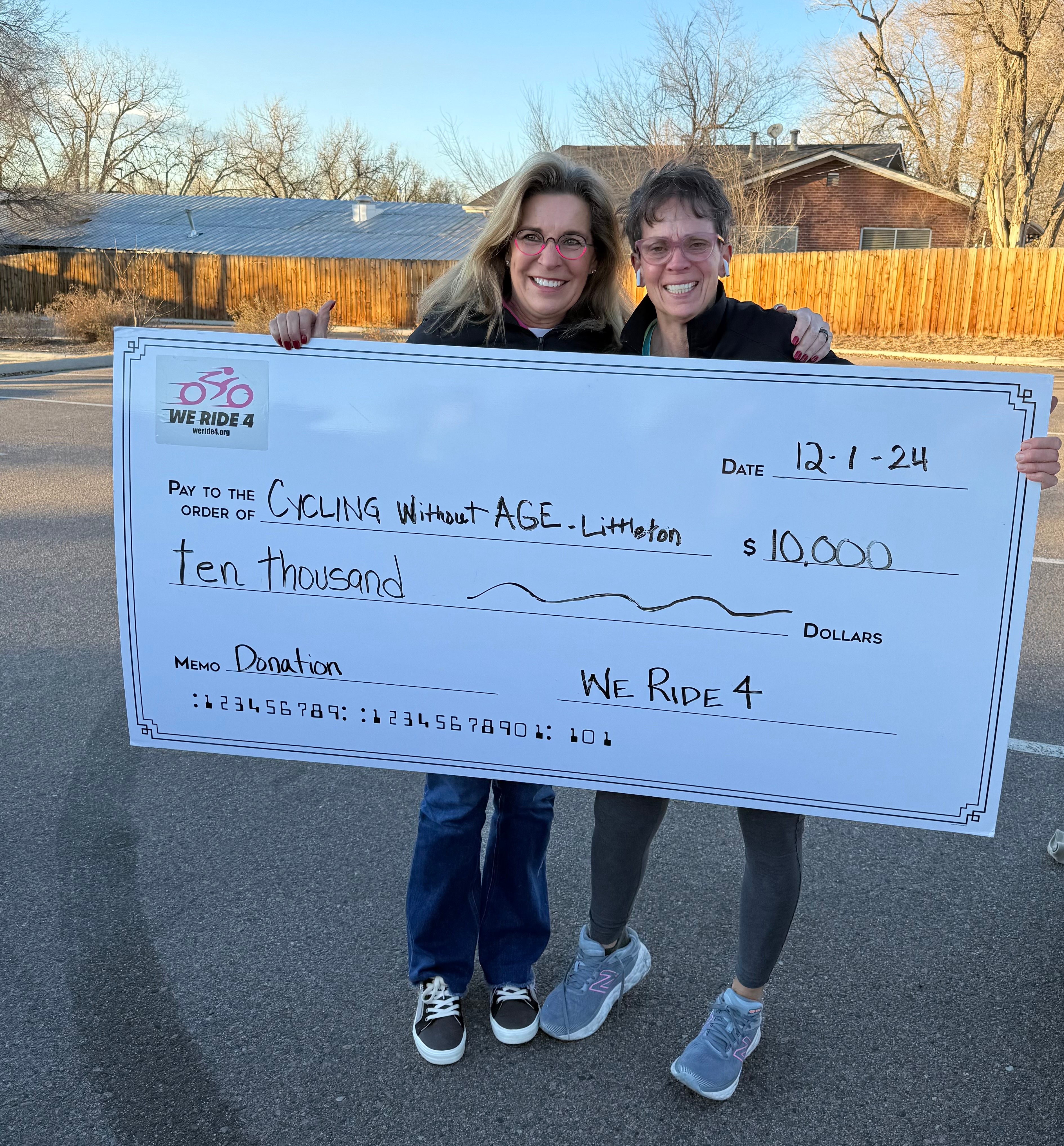 Two women holding a large check outdoors, smiling, with trees and a fence in the background.