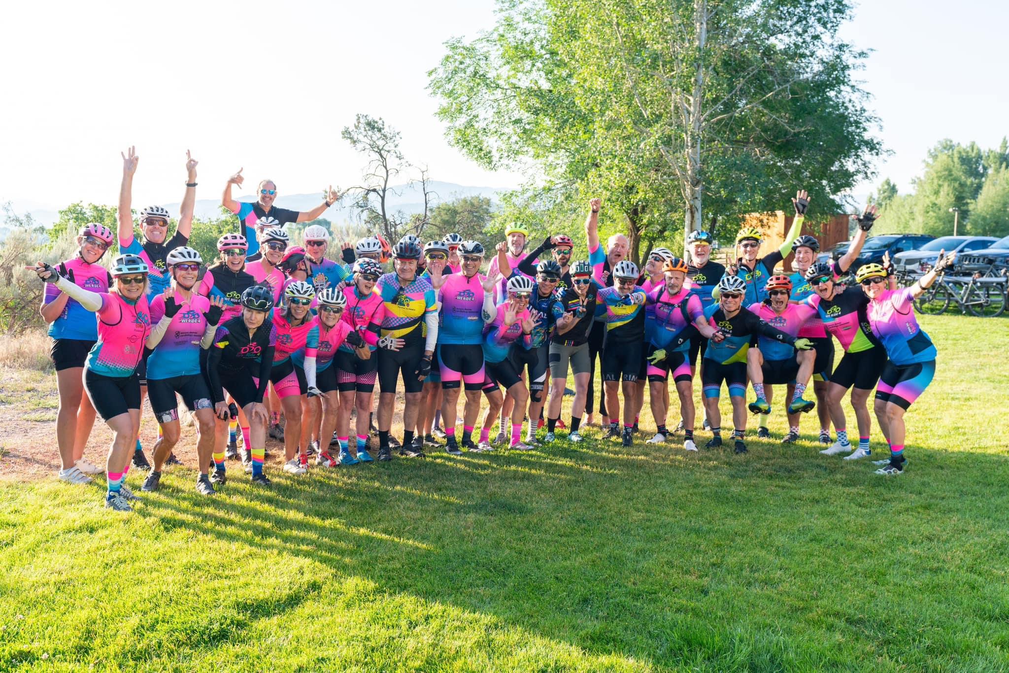 Group of people outdoors on grass, some raising arms, wearing colorful athletic clothing, trees and cars in background.