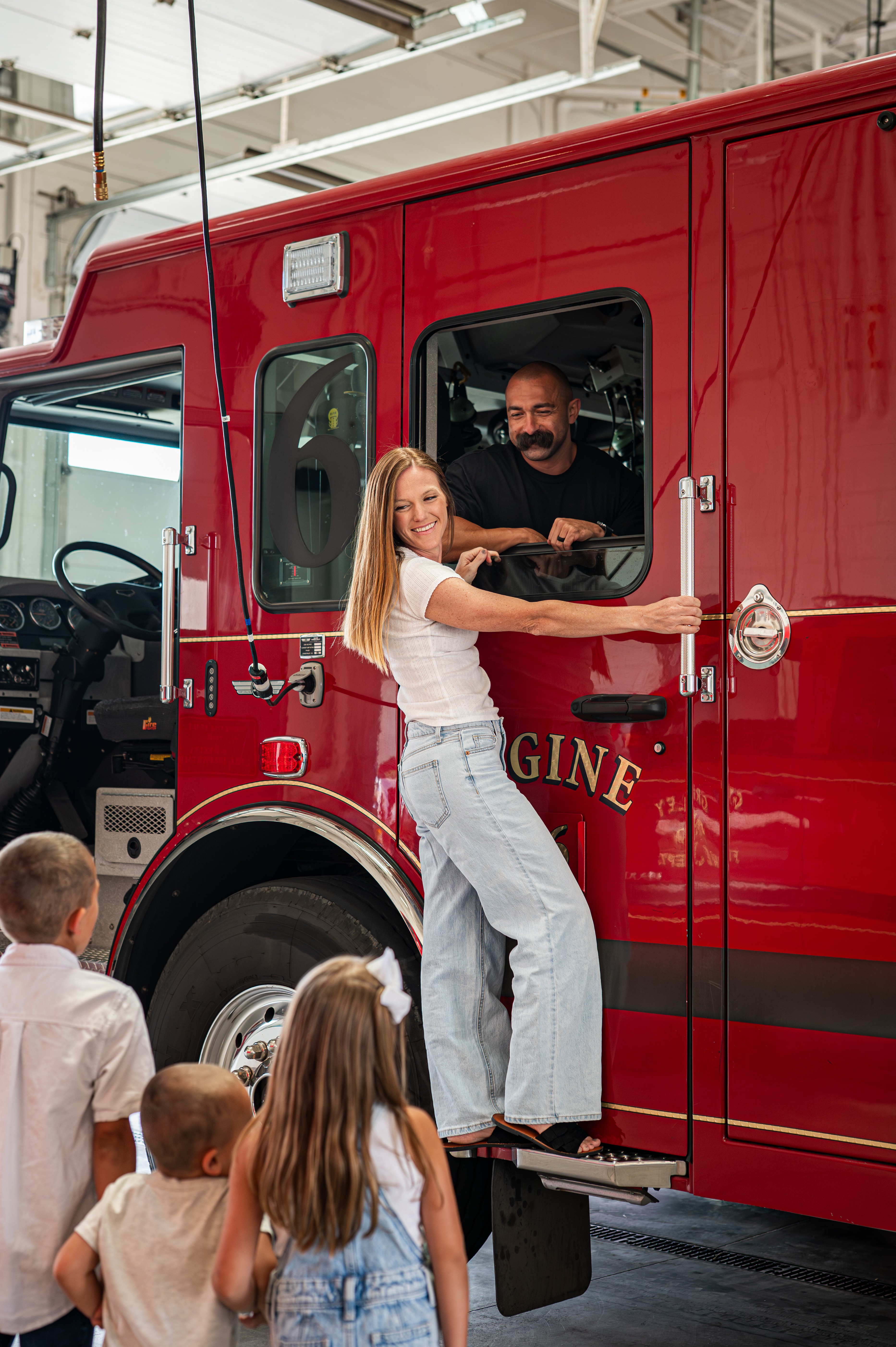 Girl hands a tray to firefighter in red fire truck, children watch inside a fire station, bright indoor setting.