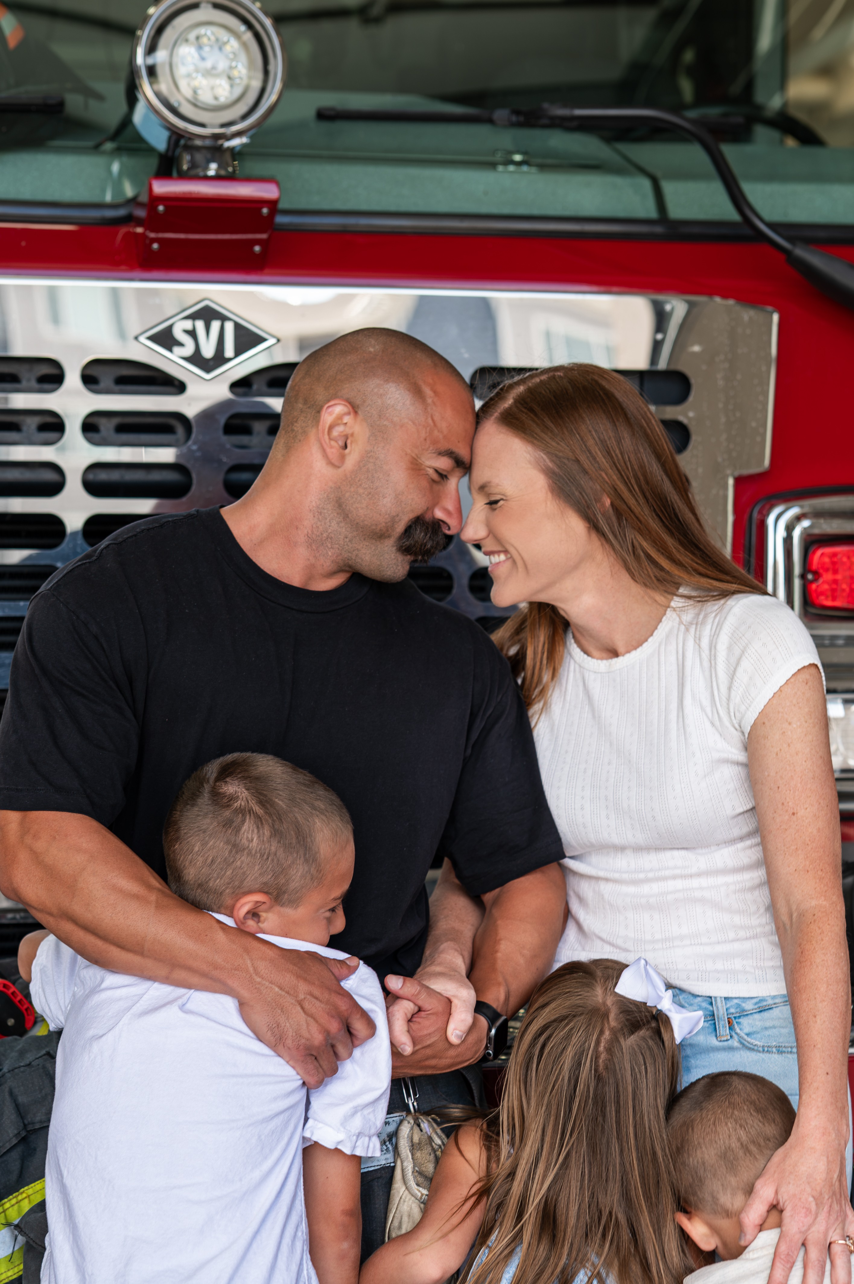 Family smiling together in front of a fire truck, with two children and two adults close and happy.