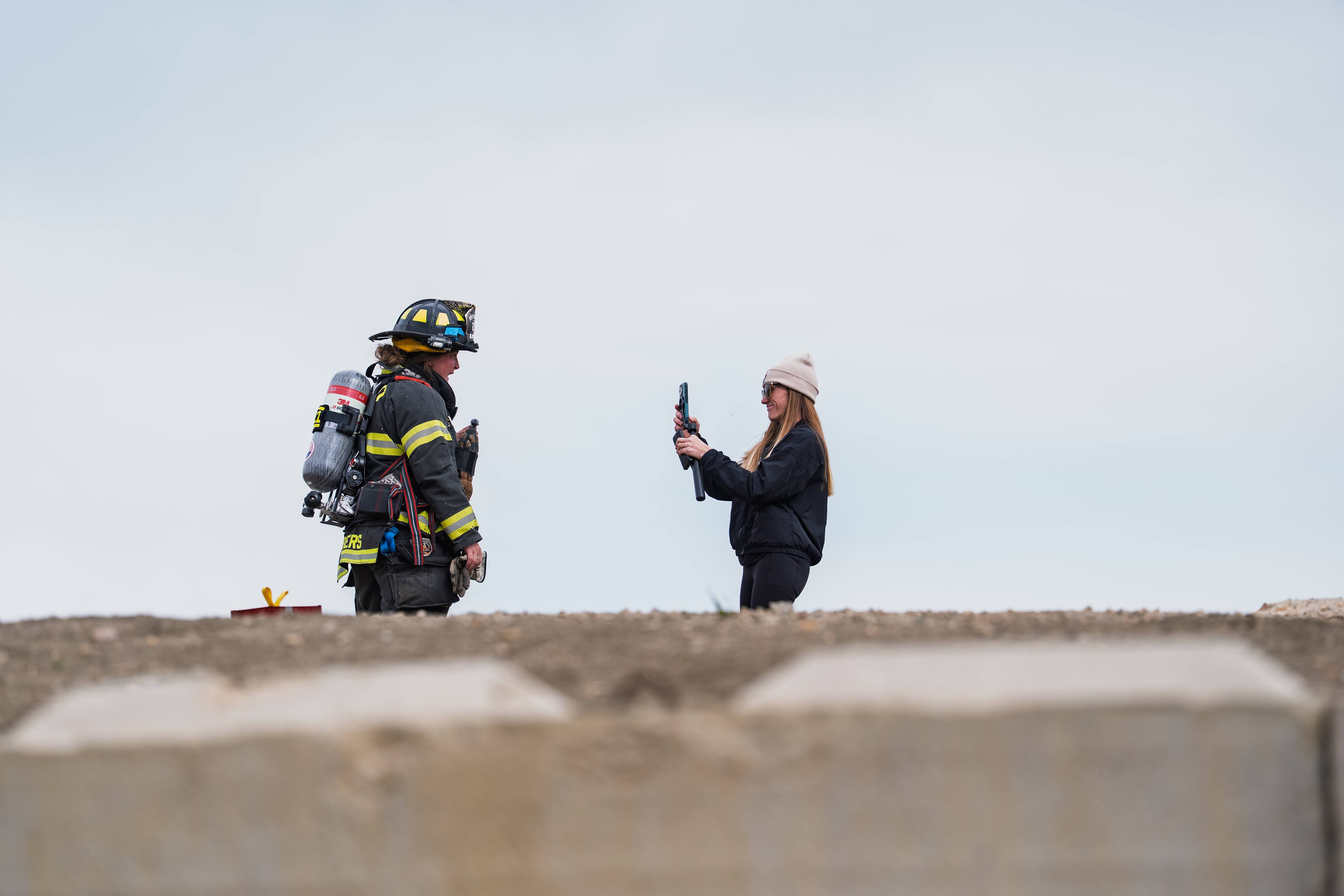 Firefighter and woman standing outdoors, woman holding a phone, sky background, ground surface visible.