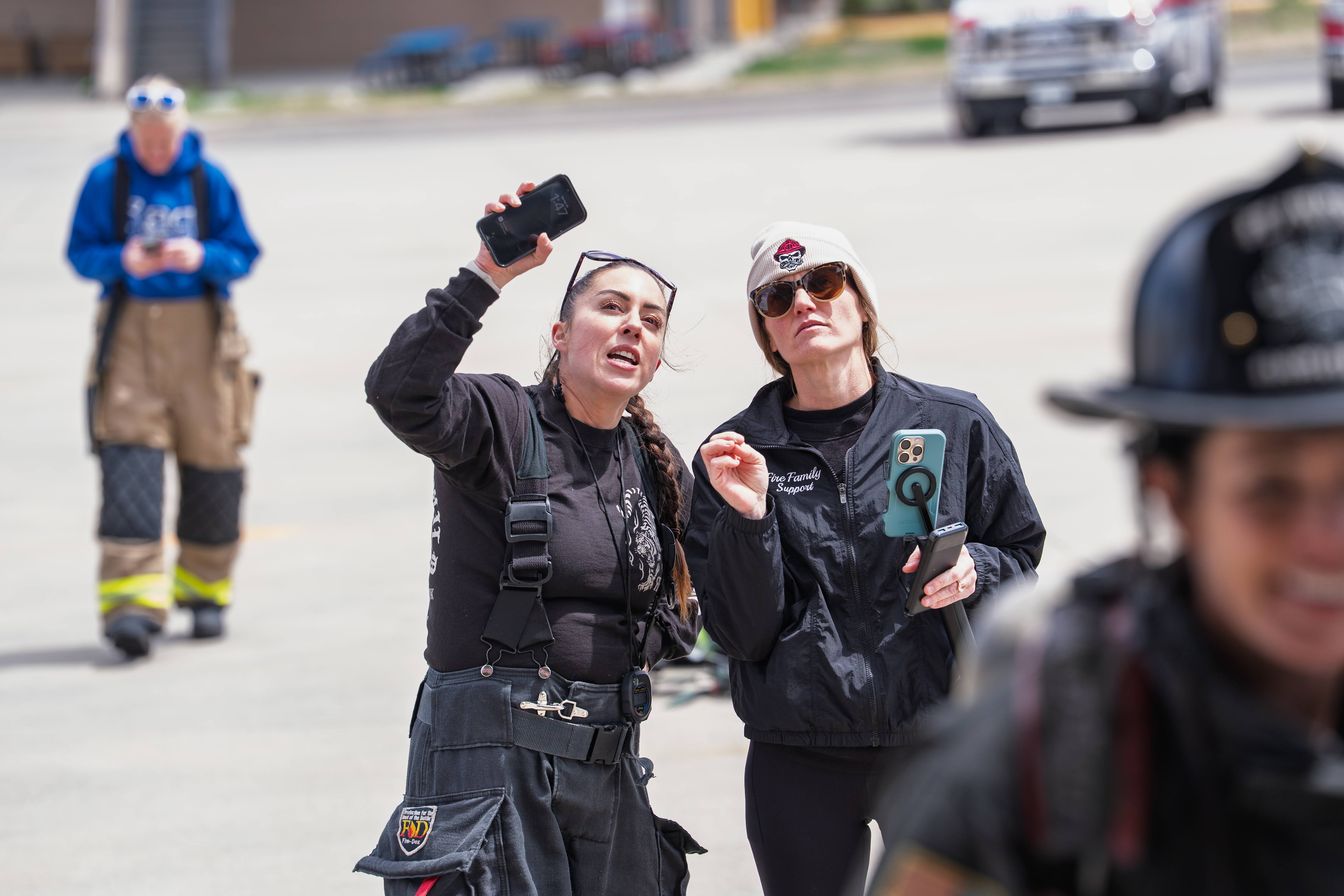 Two women in uniform talking outdoors, one holding a phone, with a person in uniform in the foreground and a person in the background.