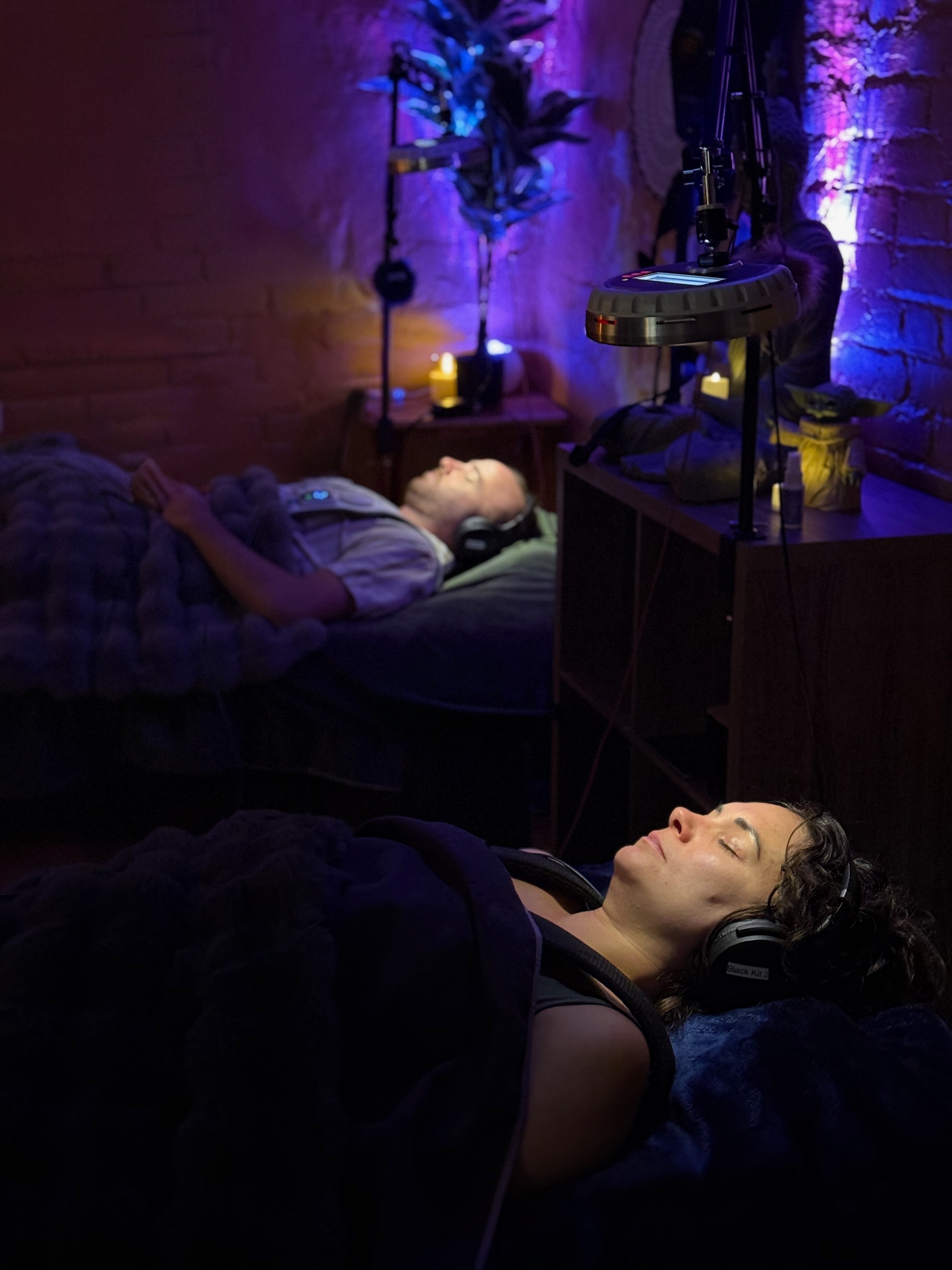 Two women lie in beds with headphones, one awake and the other sleeping, in a dimly lit room with purple lighting.