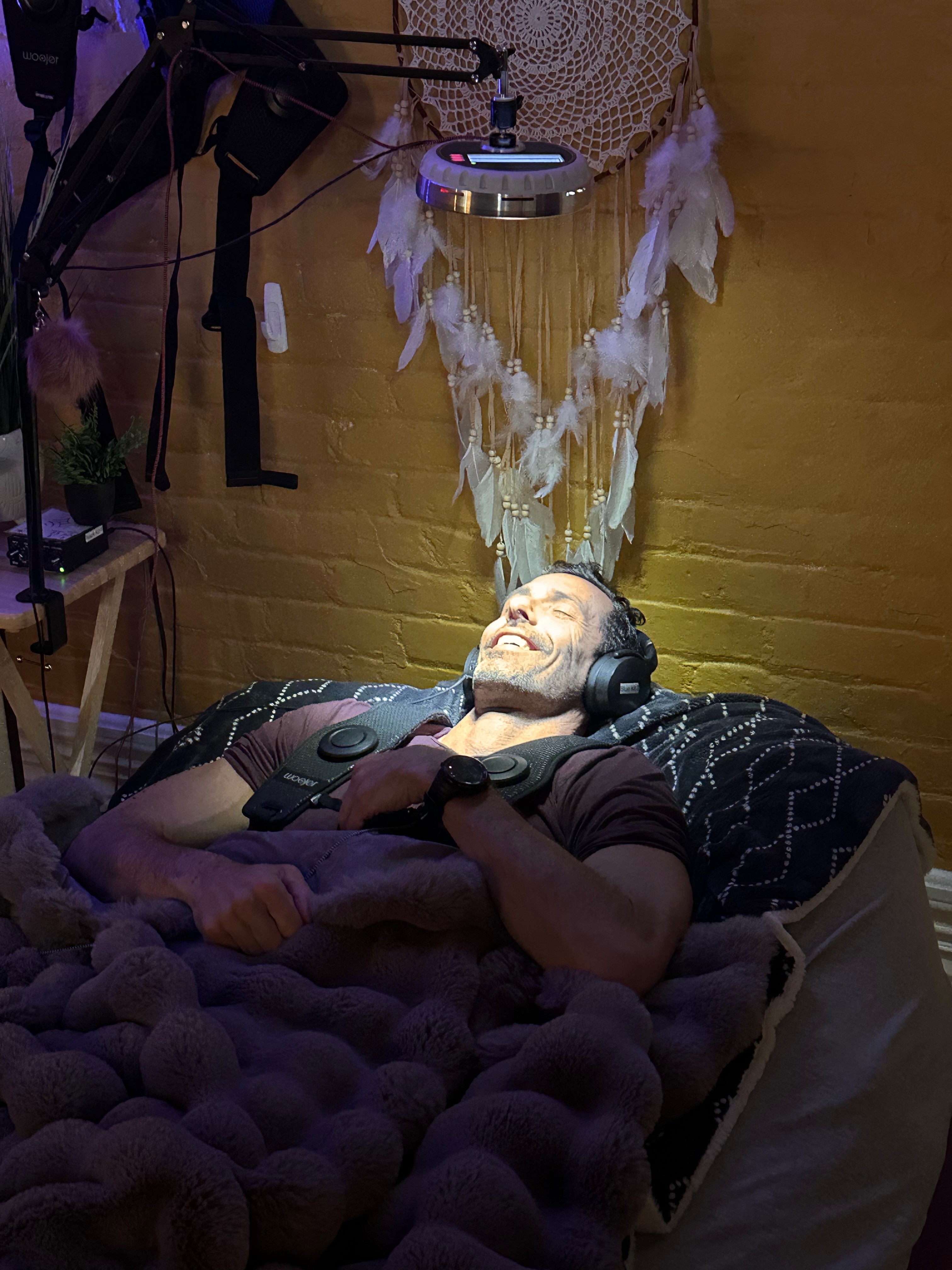 Person lying in bed with headphones, smiling, with decorative wall hanging above, in a cozy room.