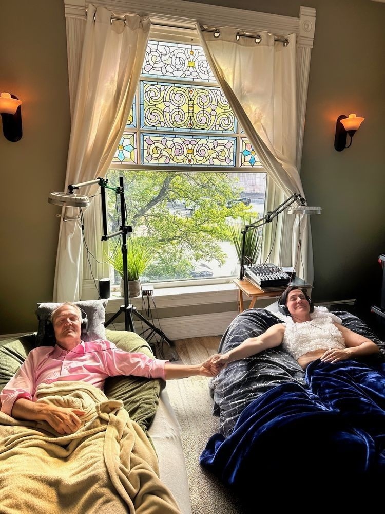 Two people lying in beds holding hands in a room with a large window, curtains, and decorative stained glass, daytime.