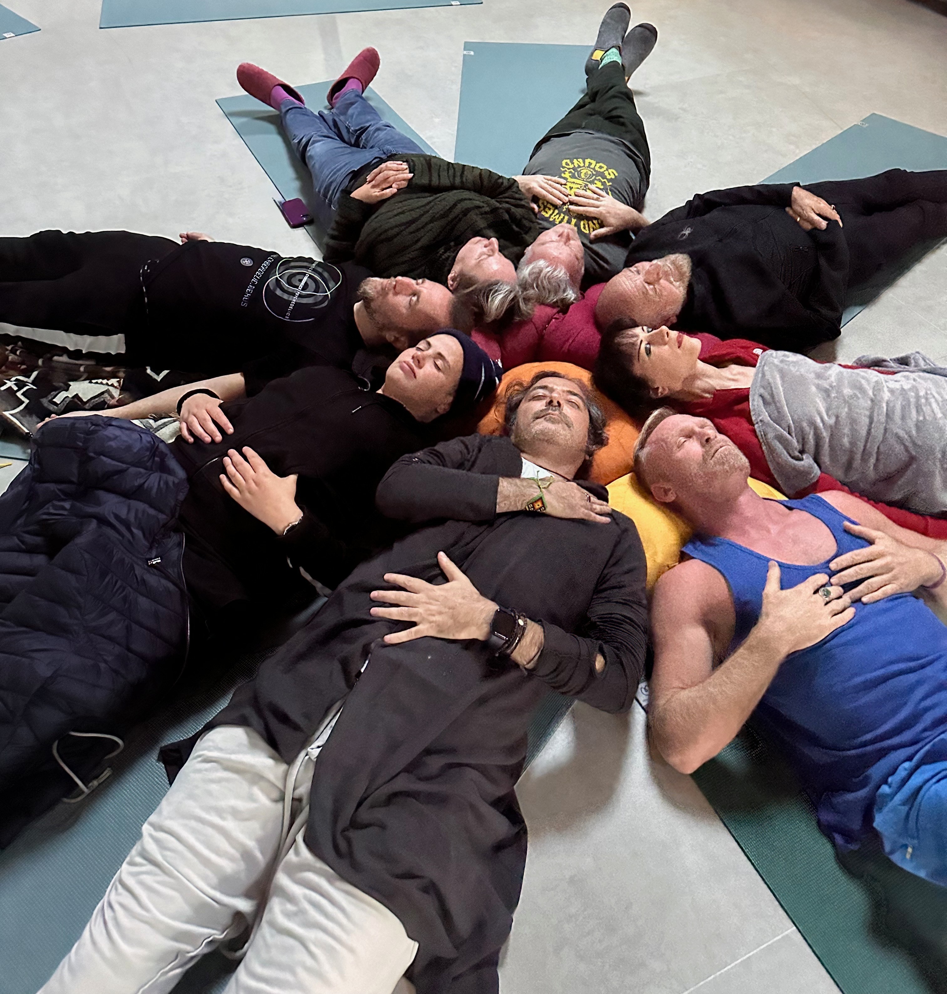 Group of people lying on the floor in a circle, heads together, eyes closed, some with hands on chest or stomach.