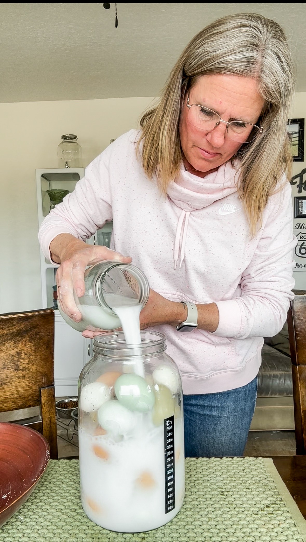 Woman pouring liquid into a jar filled with colorful eggs, standing at a table with a green woven placemat.