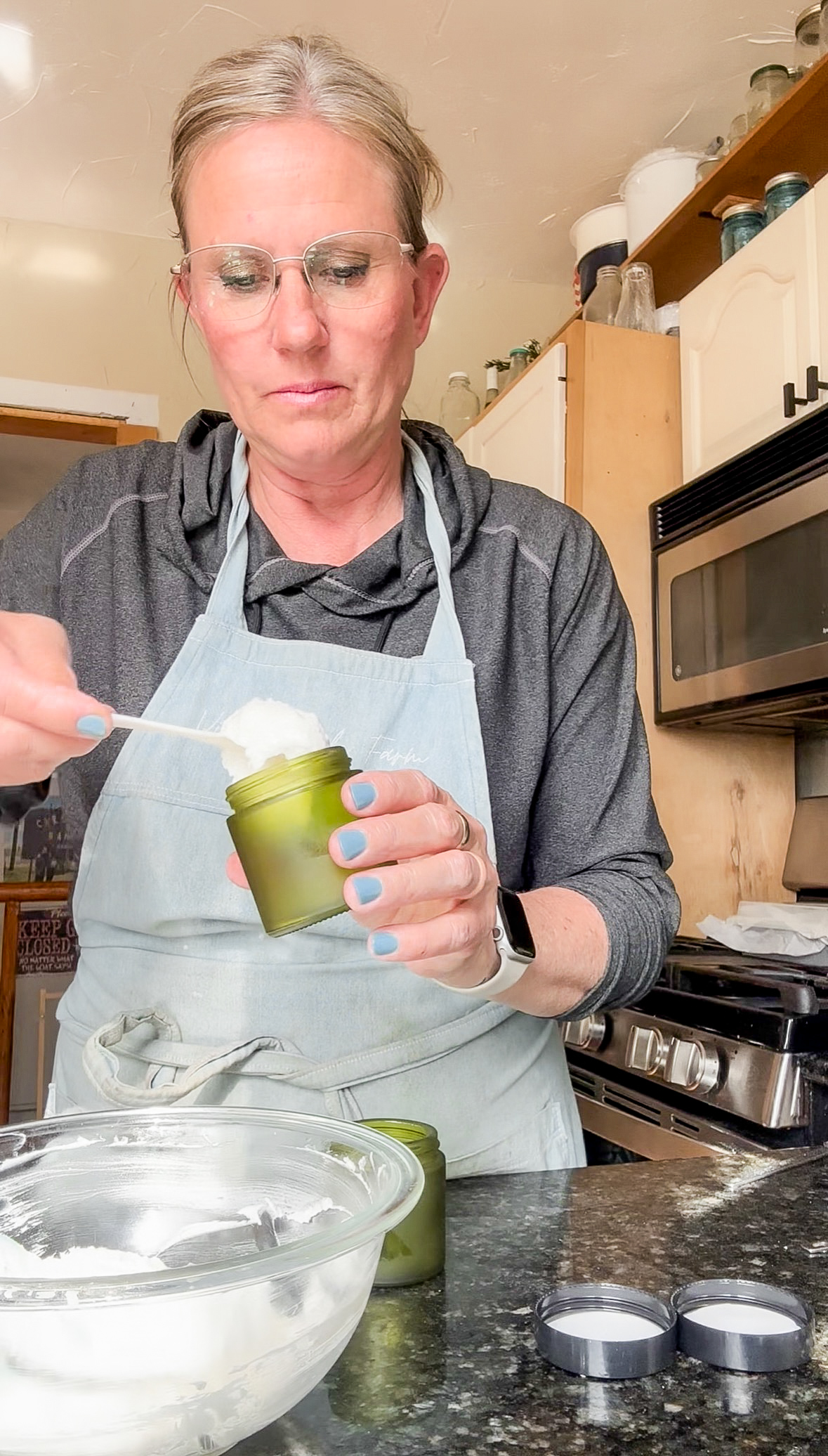 Woman in apron holding a small green container, preparing ingredients in a kitchen. Word count: 20