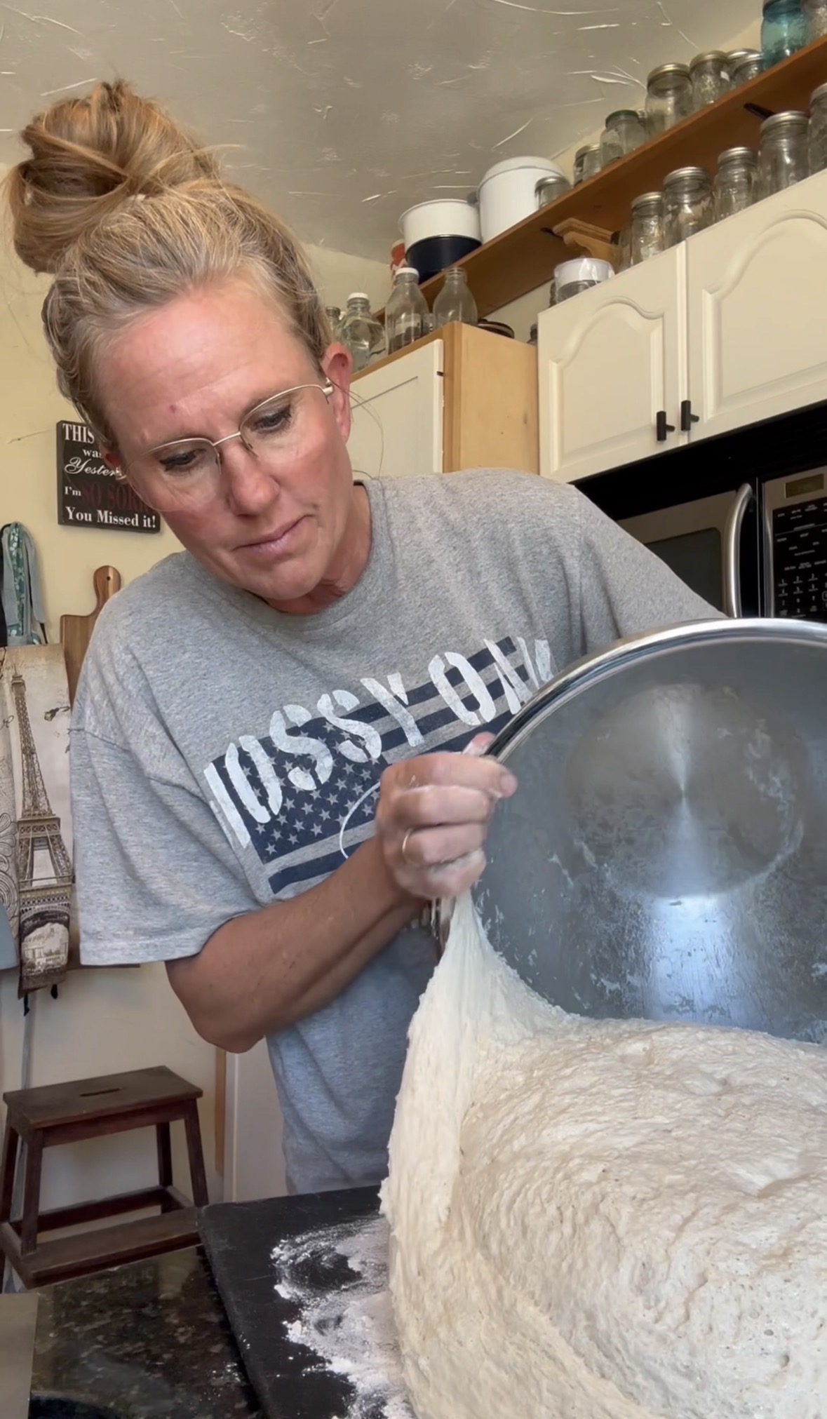 Woman pouring flour from a large metal bowl into a countertop, in a kitchen setting.