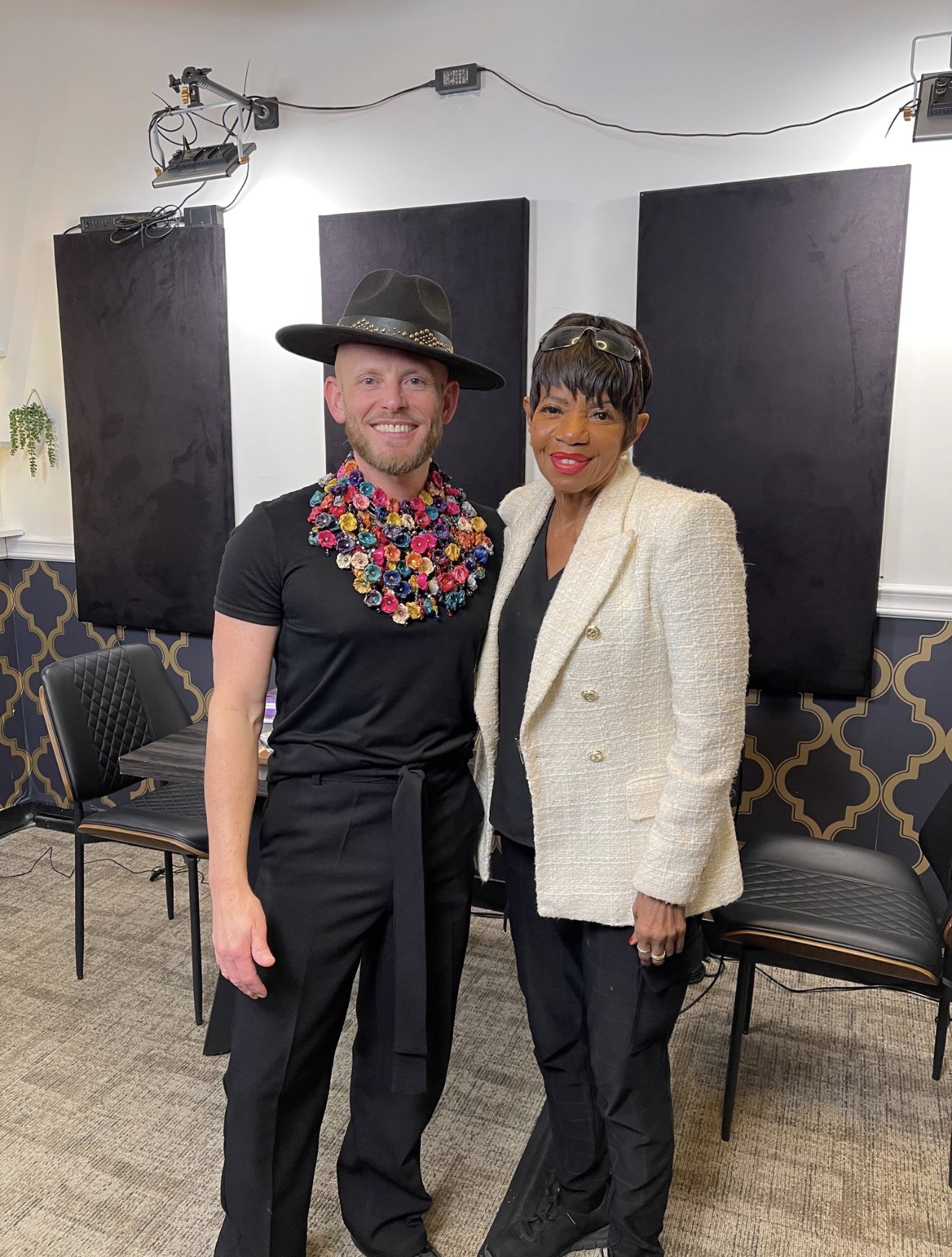 Two people standing indoors, one wearing a hat and colorful necklace, the other in a white blazer, smiling.