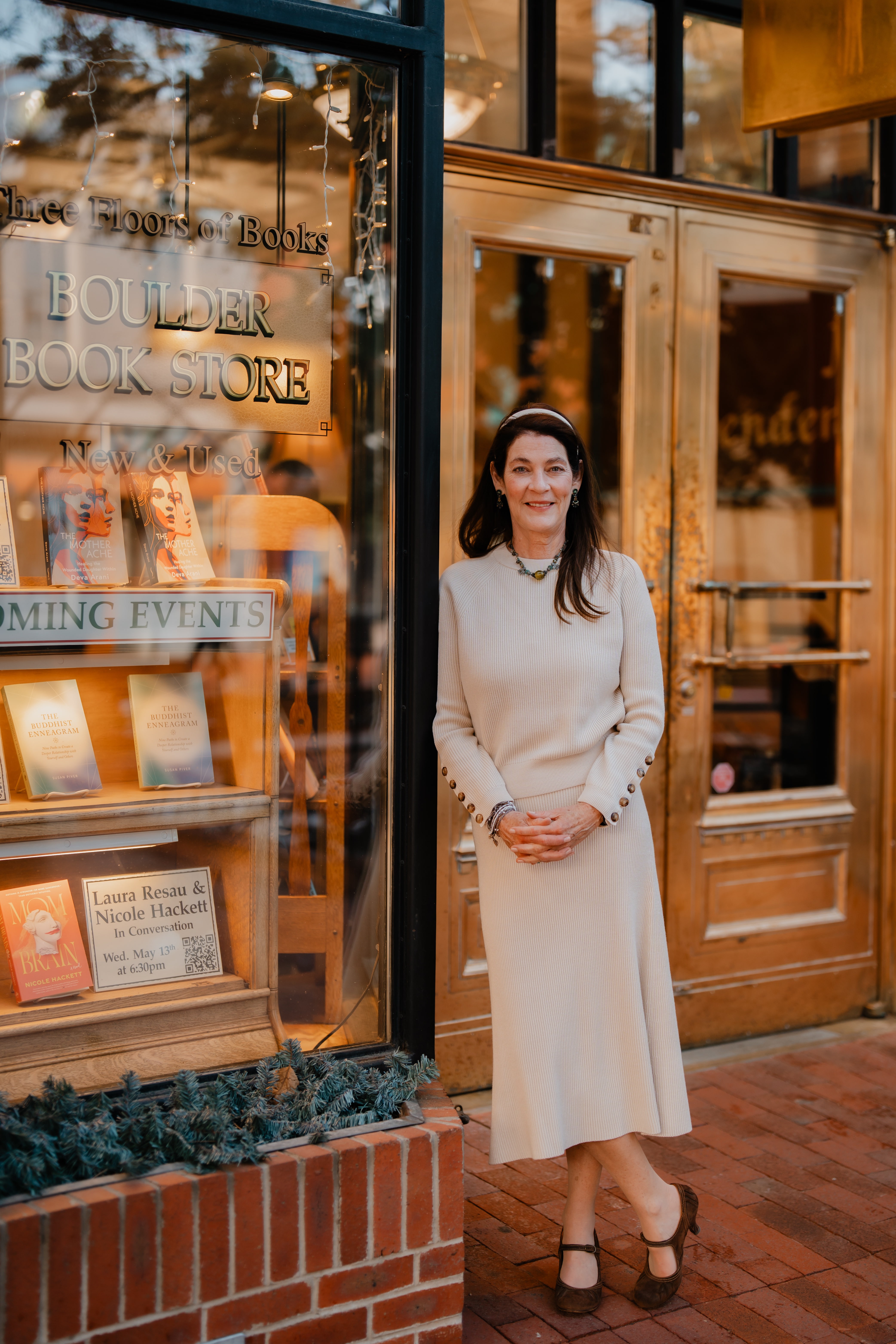 Woman in white dress standing outside bookstore with glass window and wooden door, reflected trees visible.