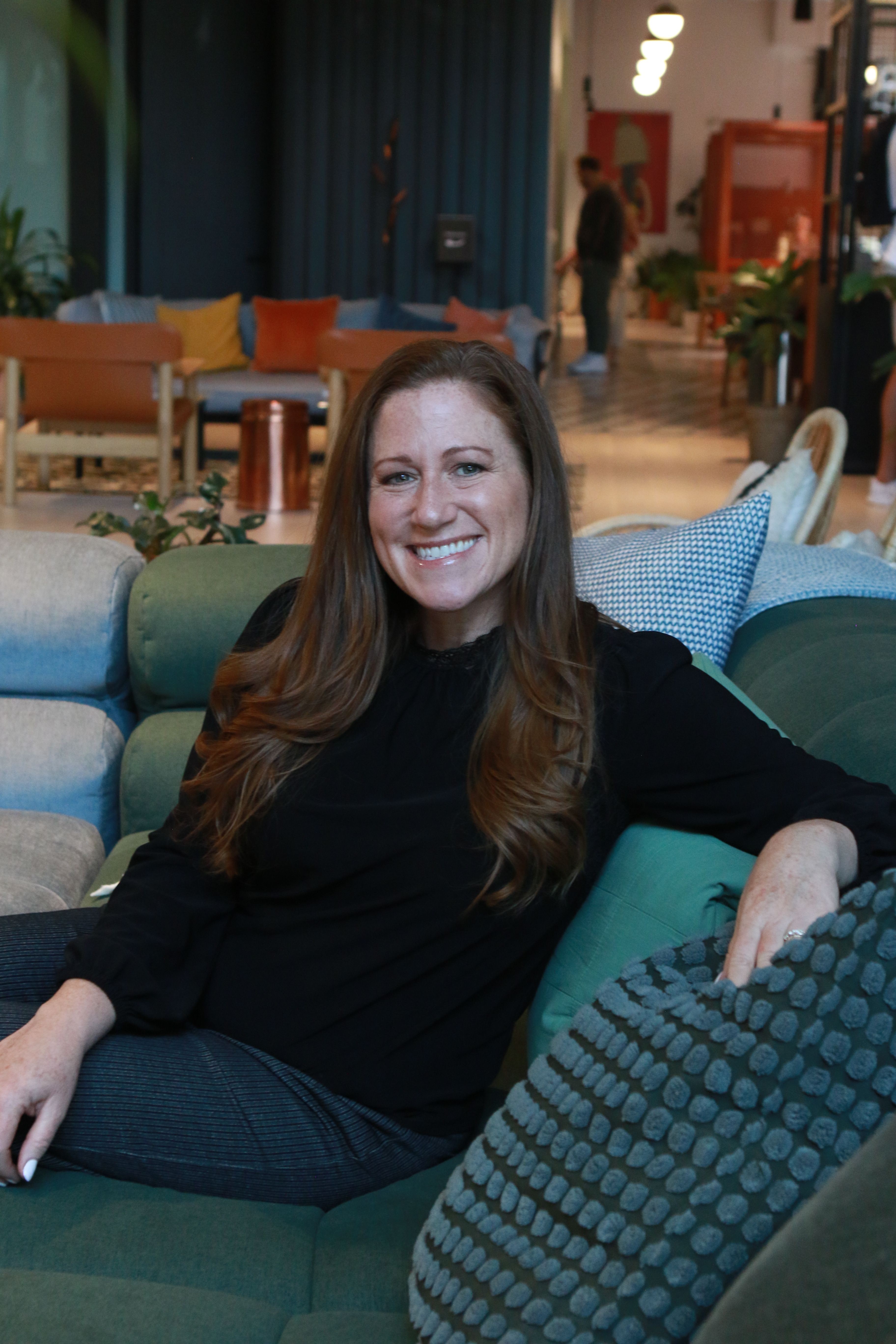 Woman with long hair sitting on a sofa, smiling, in a living room with plants and furniture.