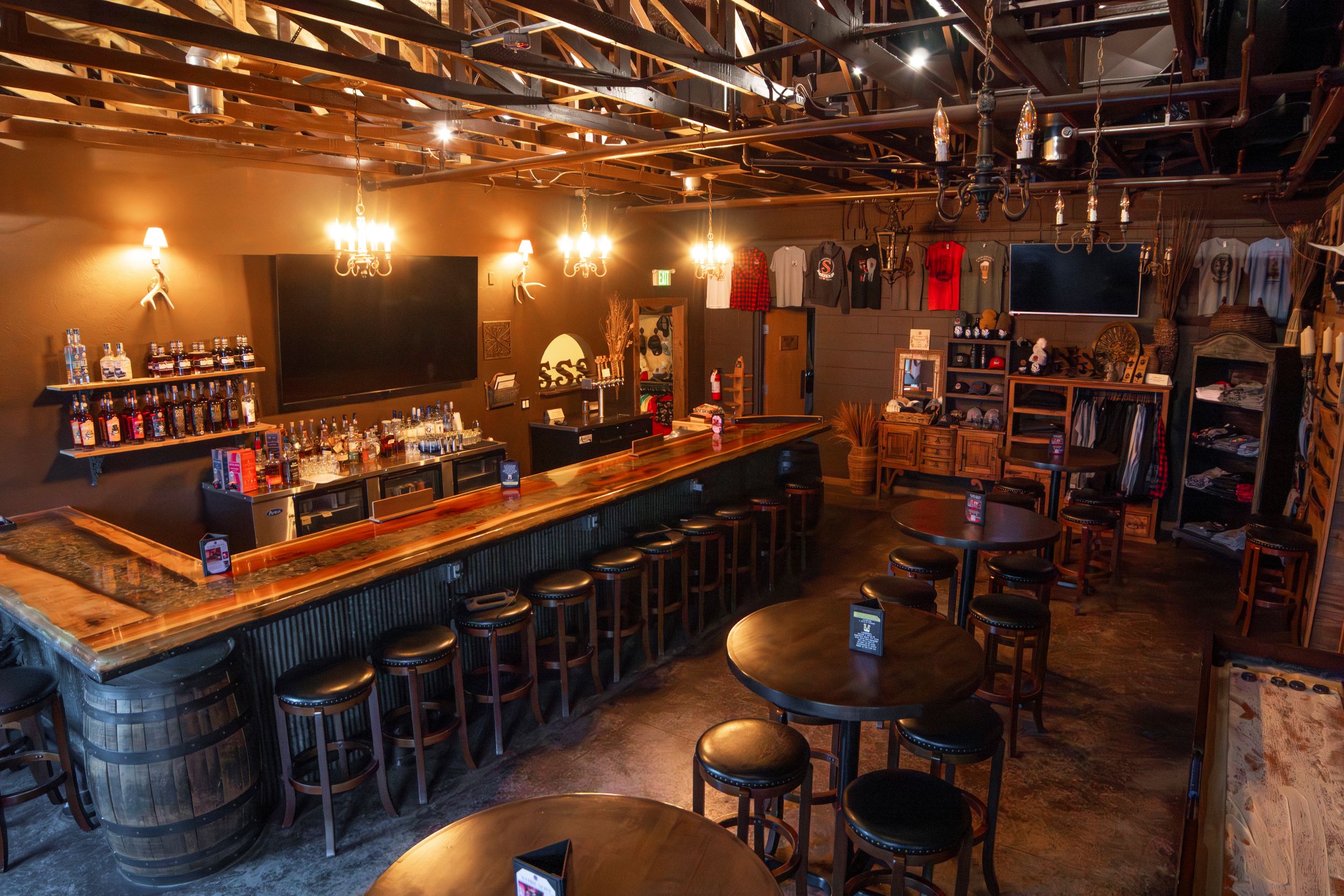 Bar with stools, shelves of bottles, and a large TV, warm lighting, and wooden decor in a cozy pub setting.