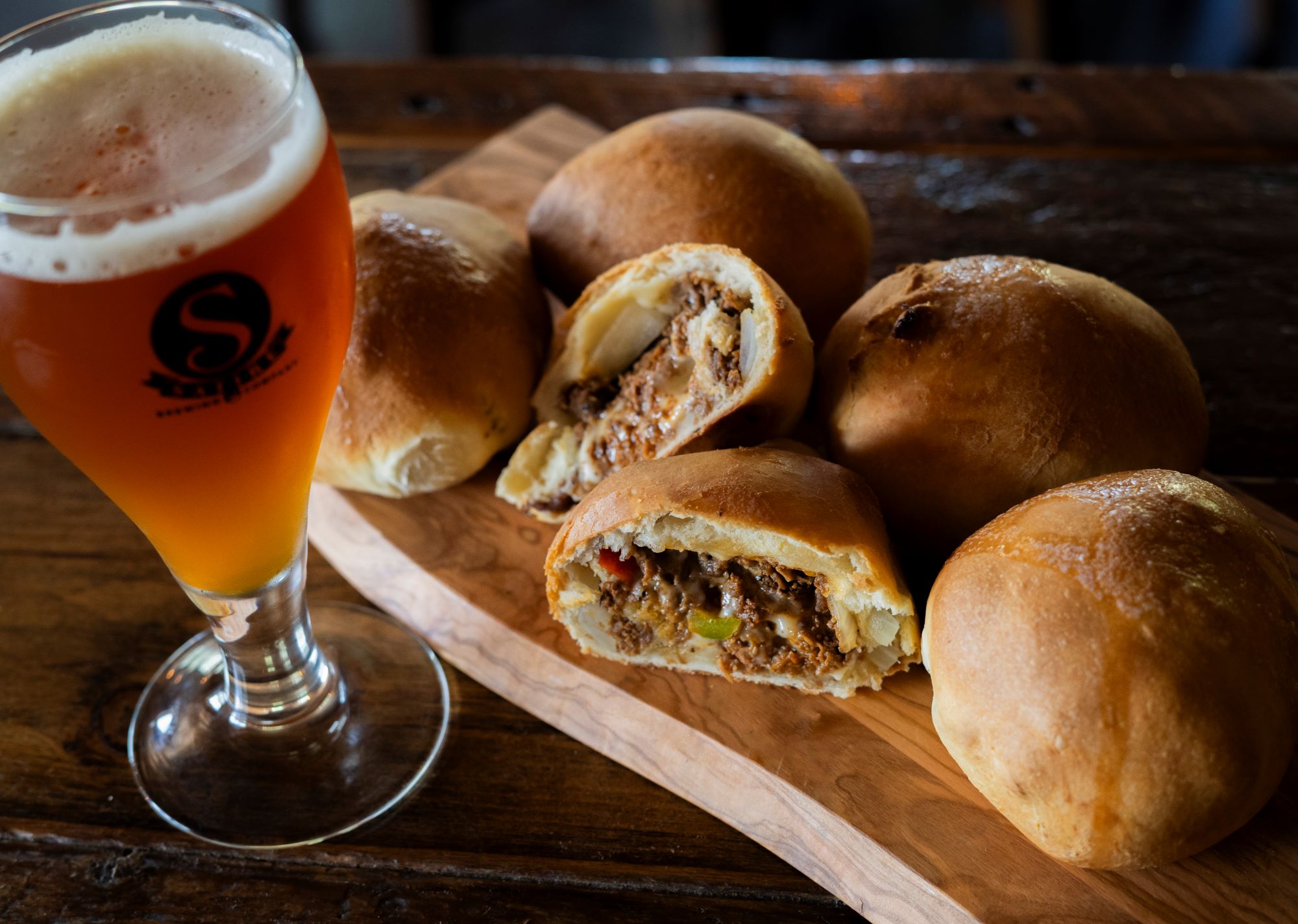 Beer in a glass and several bread rolls, some cut open showing filling, on a wooden surface.
