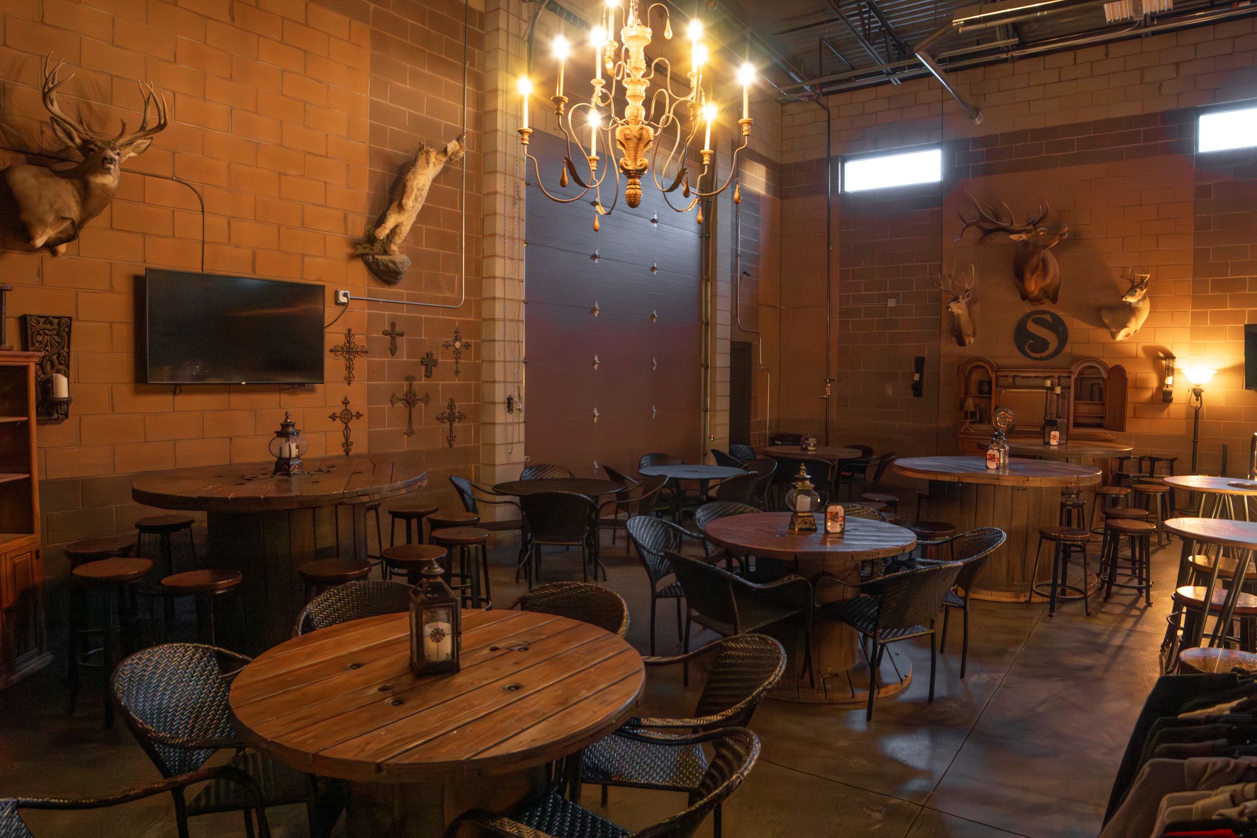 Interior of a rustic restaurant with wooden tables, chairs, mounted animal heads, a chandelier, and warm lighting.