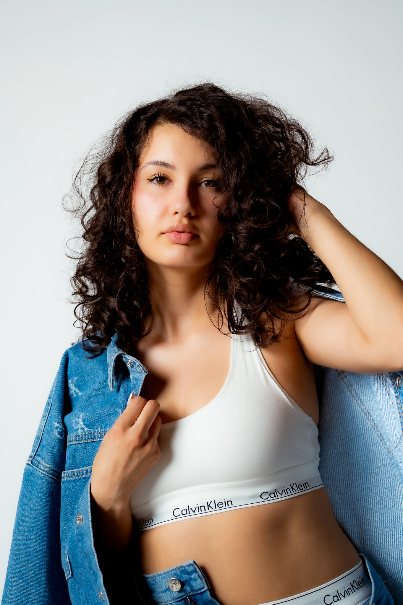 Young woman with curly hair wearing a white Calvin Klein sports bra and denim jacket, posing with hand in hair, neutral expression.