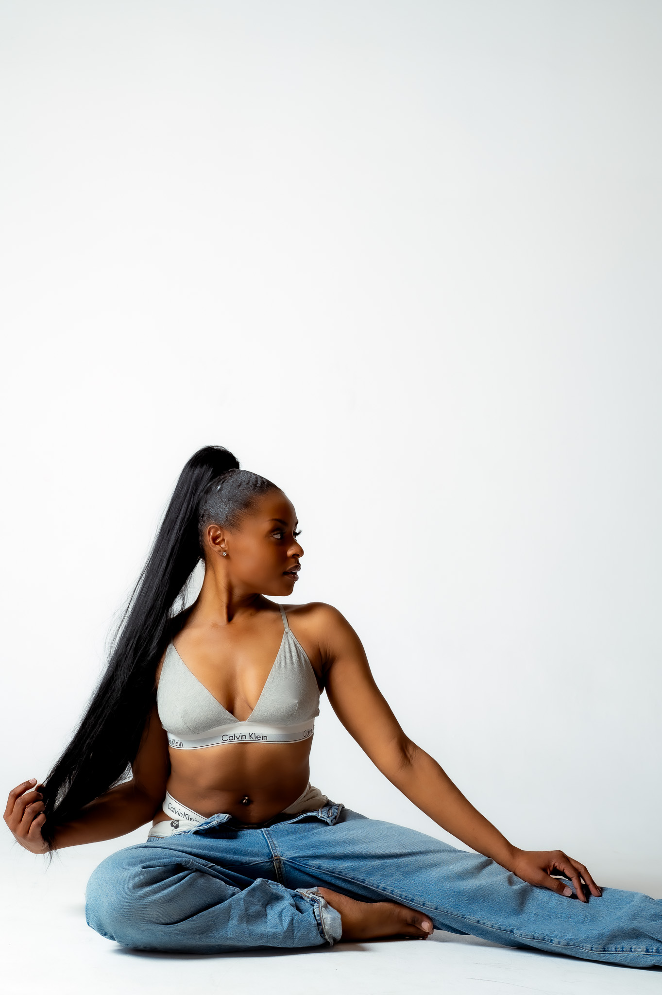 Woman with long hair sitting cross-legged on the floor, wearing a light-colored top and jeans, against a plain white background.