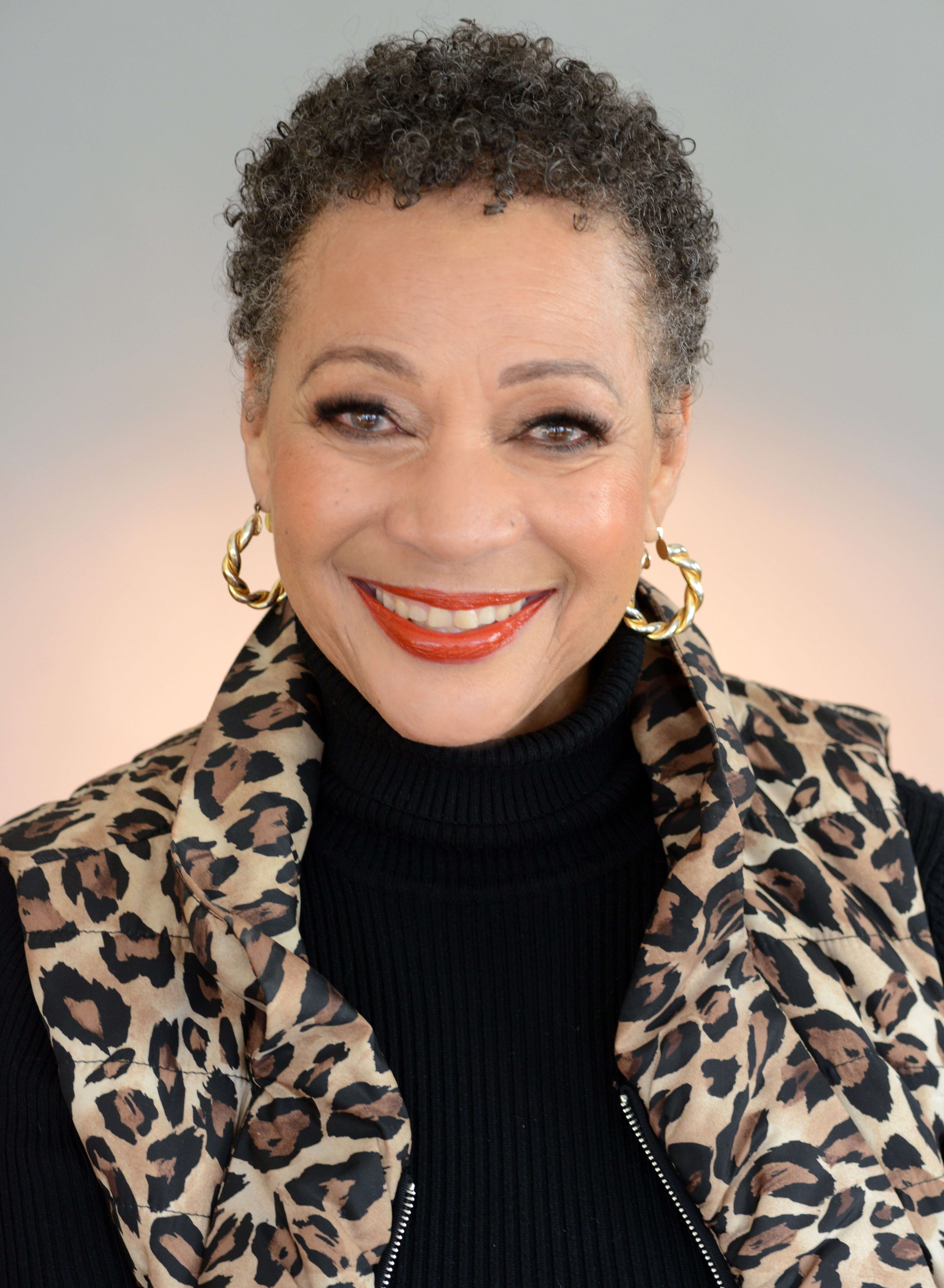 Smiling woman with short curly hair, wearing a leopard print jacket, black turtleneck, and gold hoop earrings.